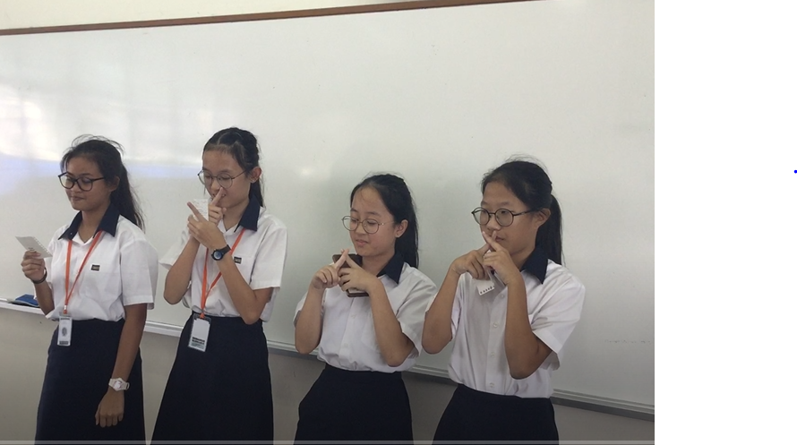 Four female students in white uniforms stand in front of a whiteboard, gesturing with their hands.