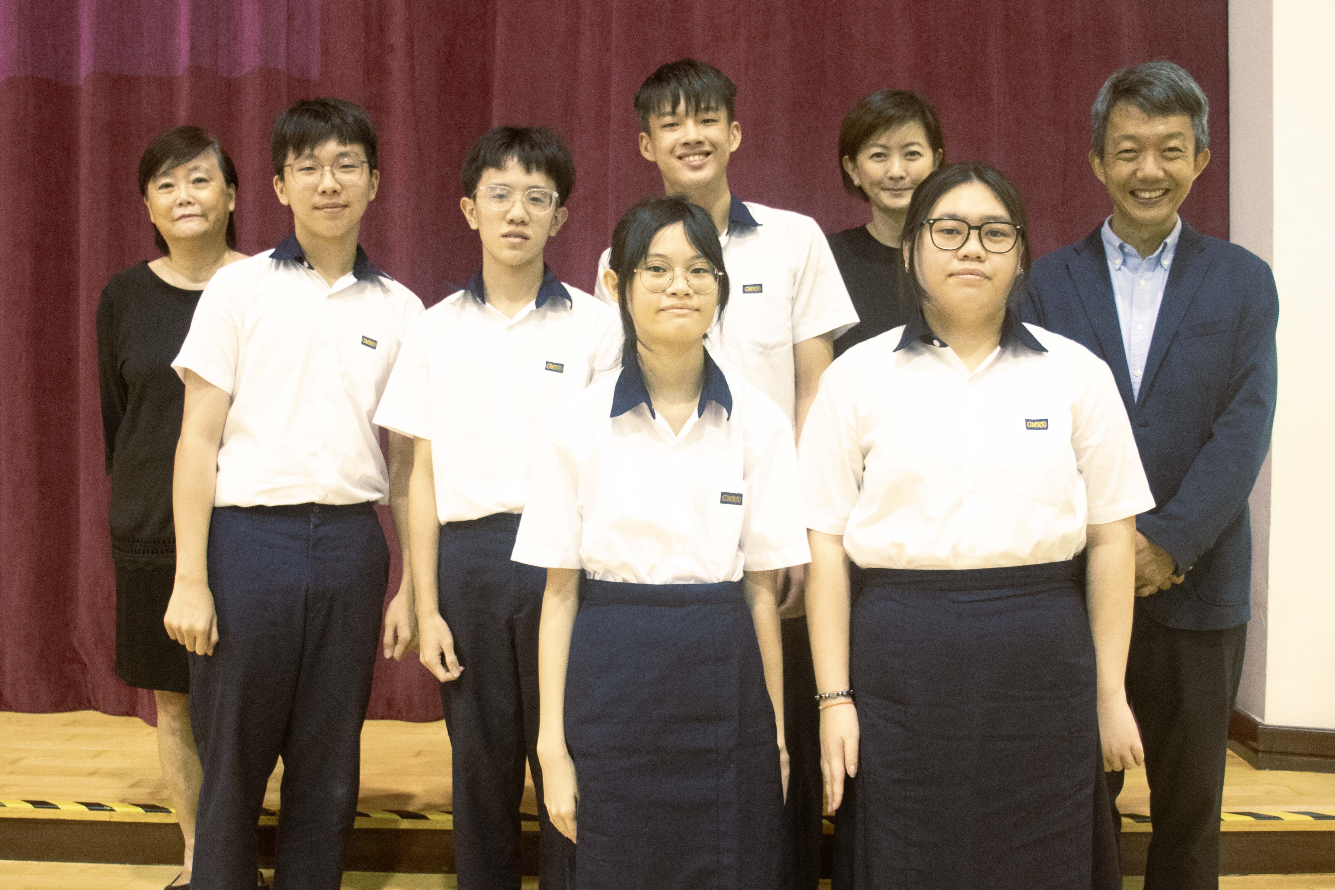 Five students in white and navy uniforms pose with two adults in front of a dark red curtain.