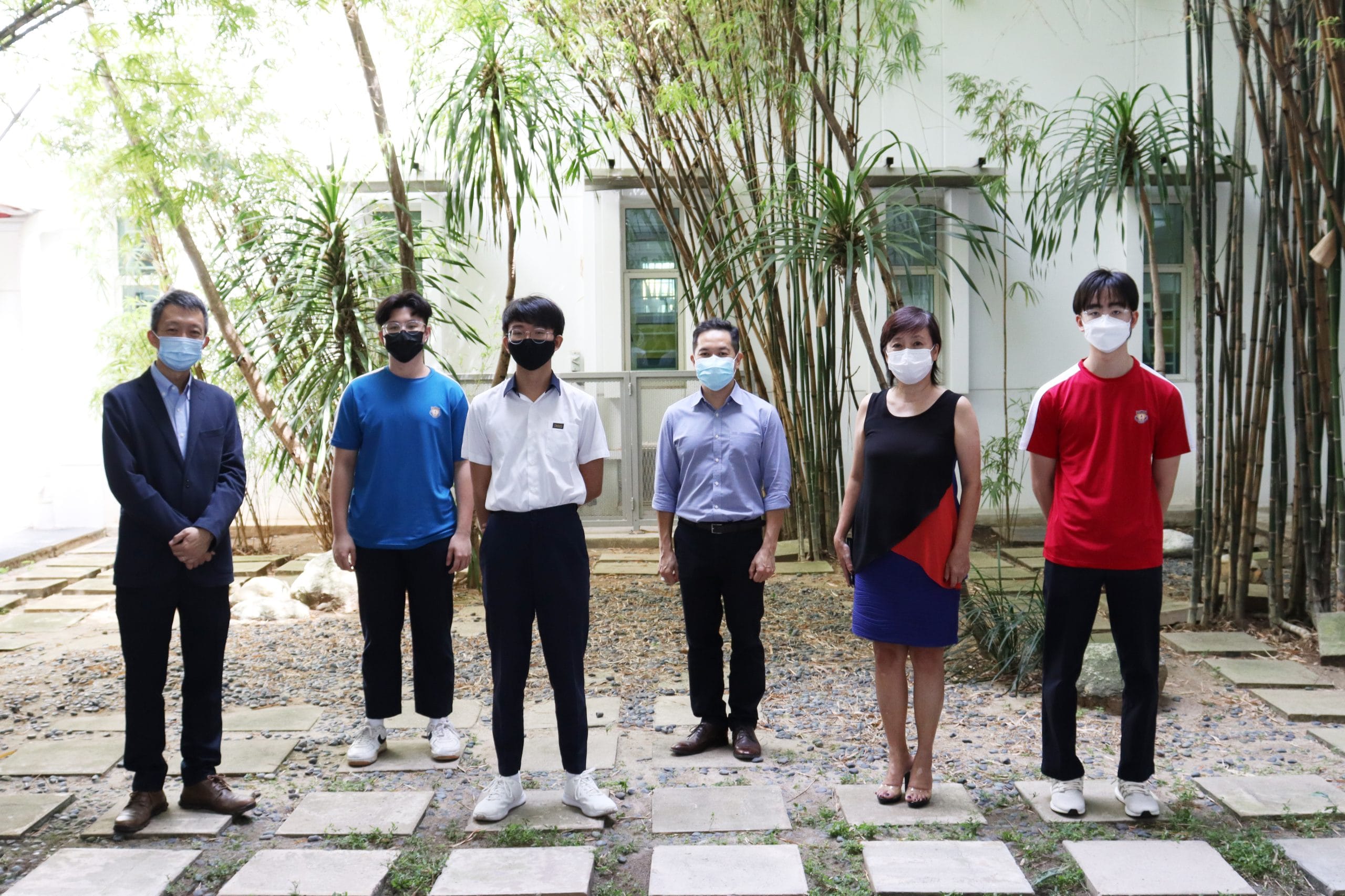 Six people wearing face masks stand on a stone-tiled courtyard with bamboo trees and a white building behind them.