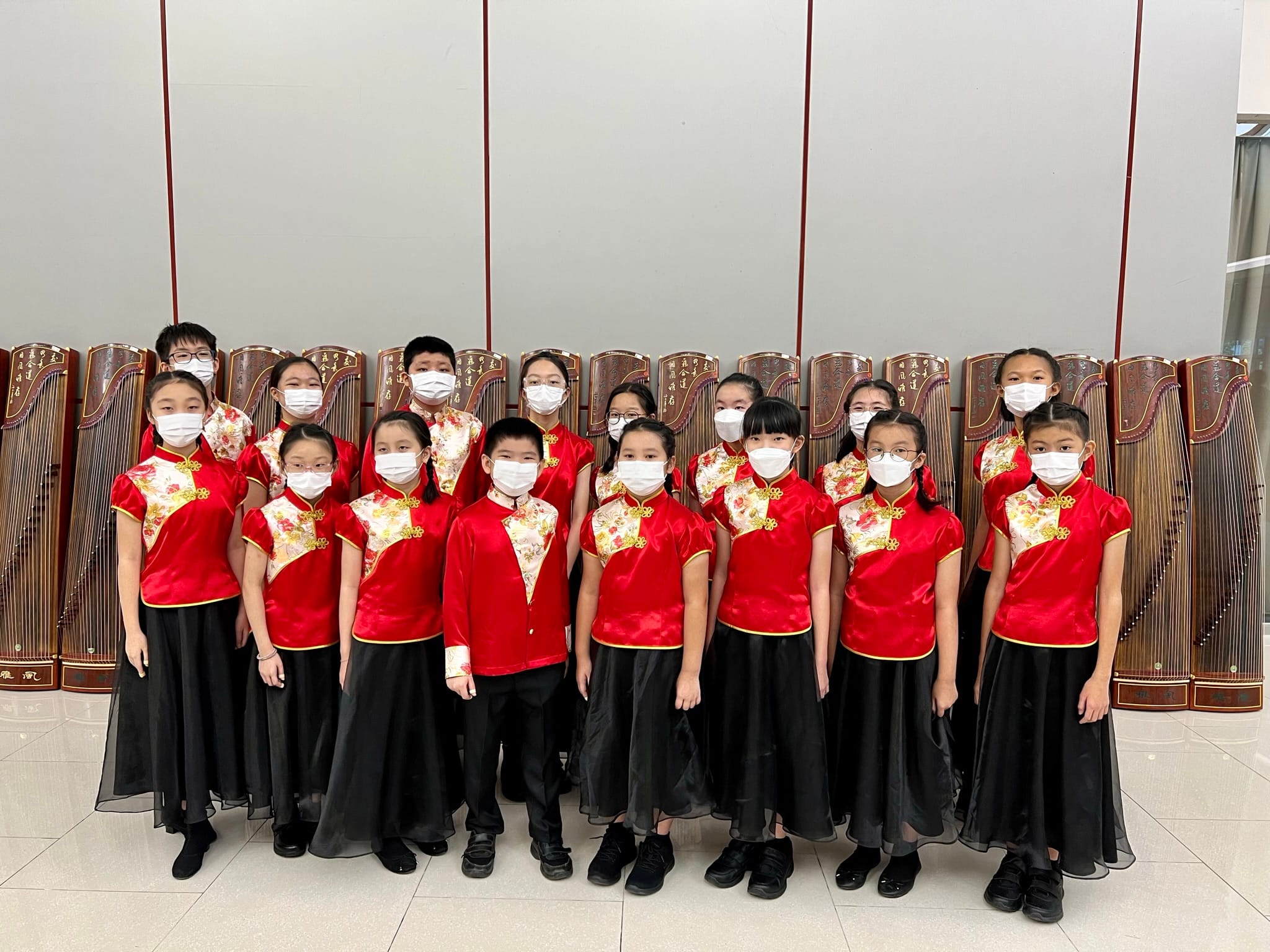 A group of children in red traditional Chinese outfits and white masks pose in front of several guzheng instruments.