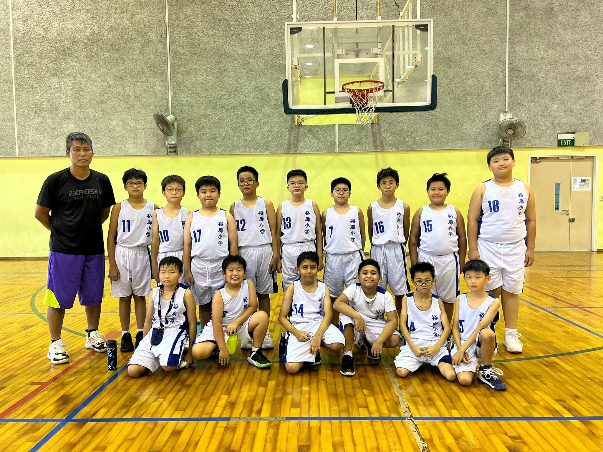 A youth basketball team in white uniforms poses with a coach on an indoor court with a hoop visible behind them.
