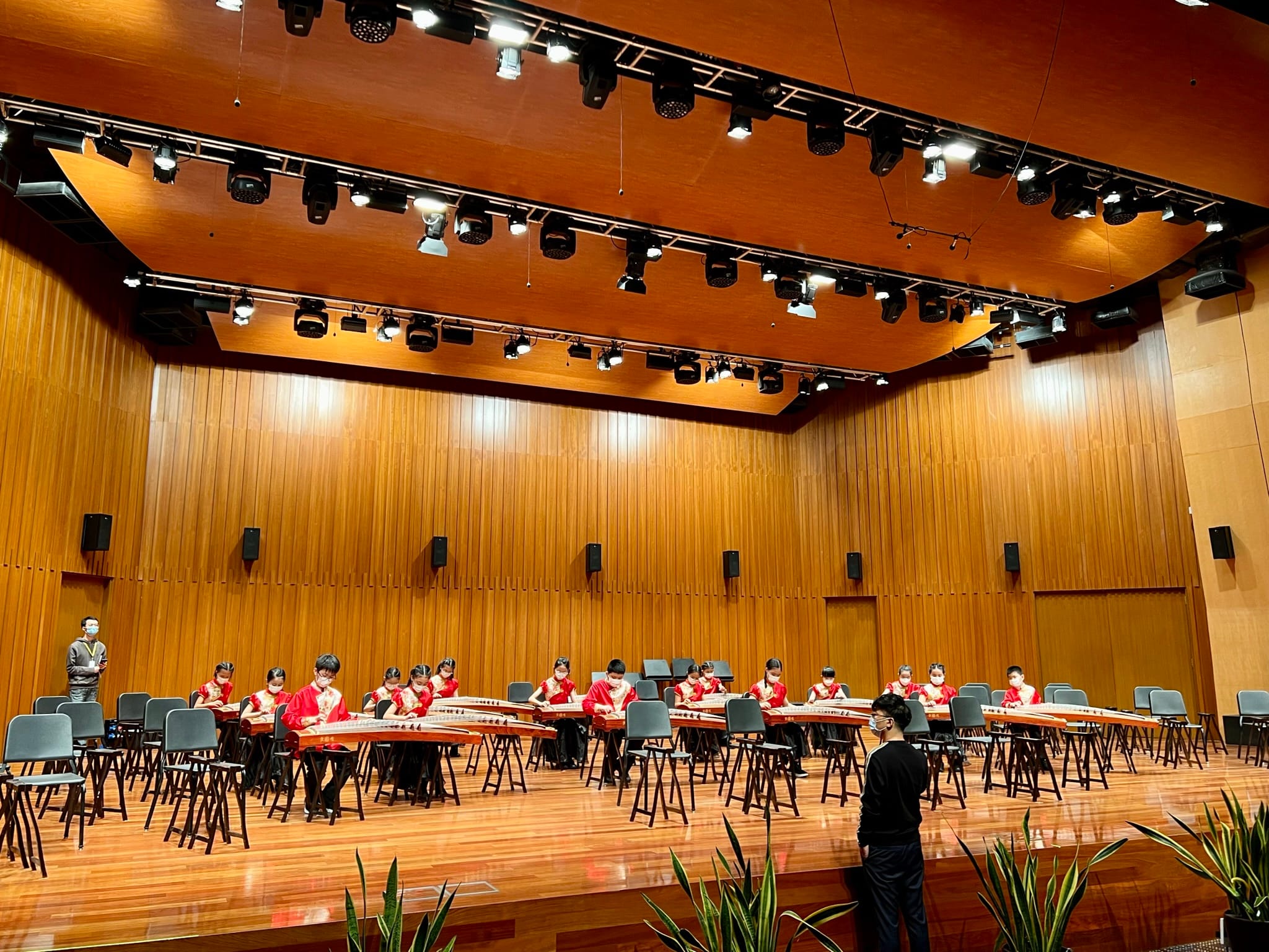 Students in red outfits play guzheng instruments on a wood-paneled concert hall stage under stage lighting.