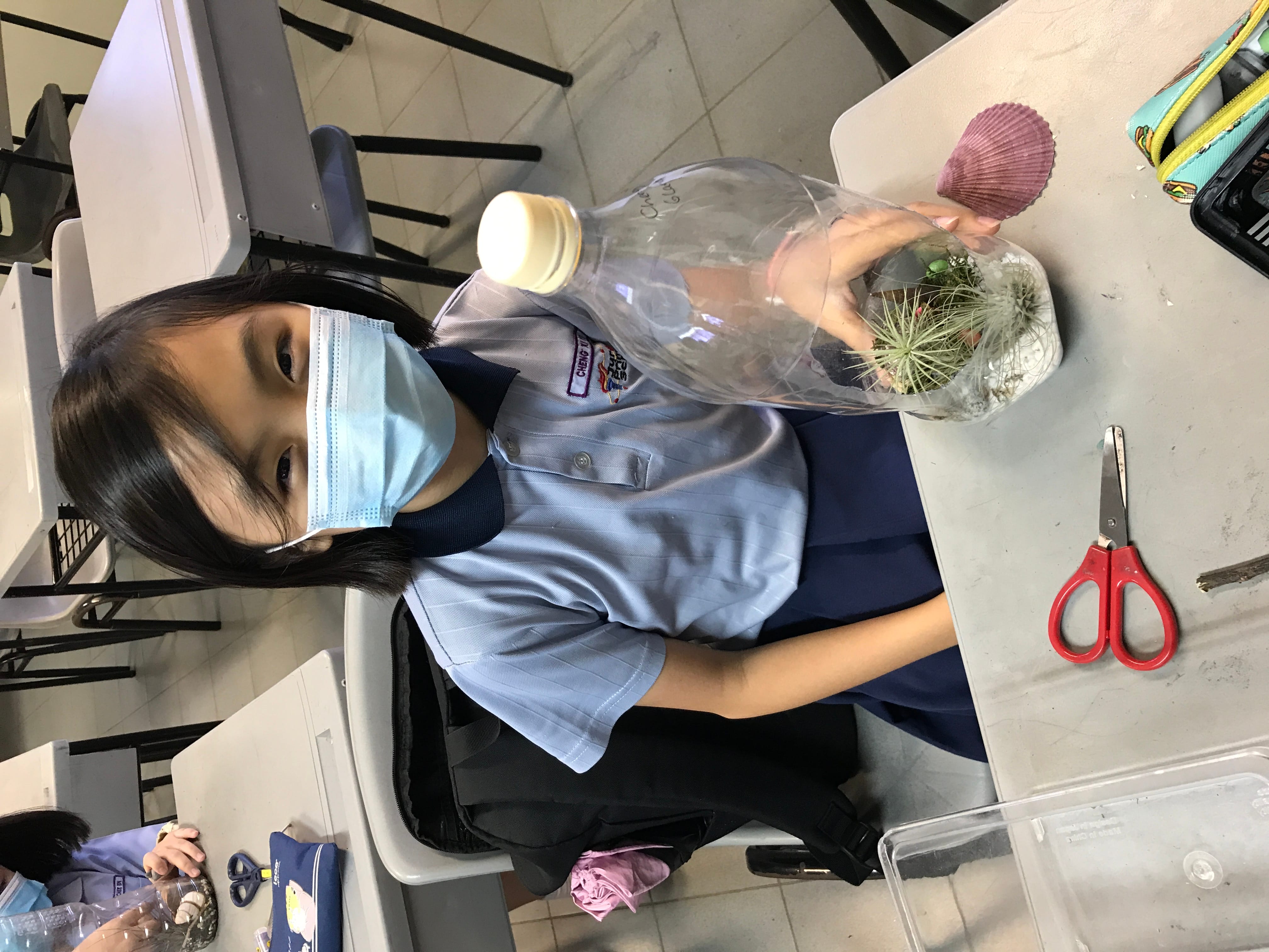 A masked schoolgirl in uniform holds a plastic bottle terrarium with small plants inside, sitting at a classroom desk.