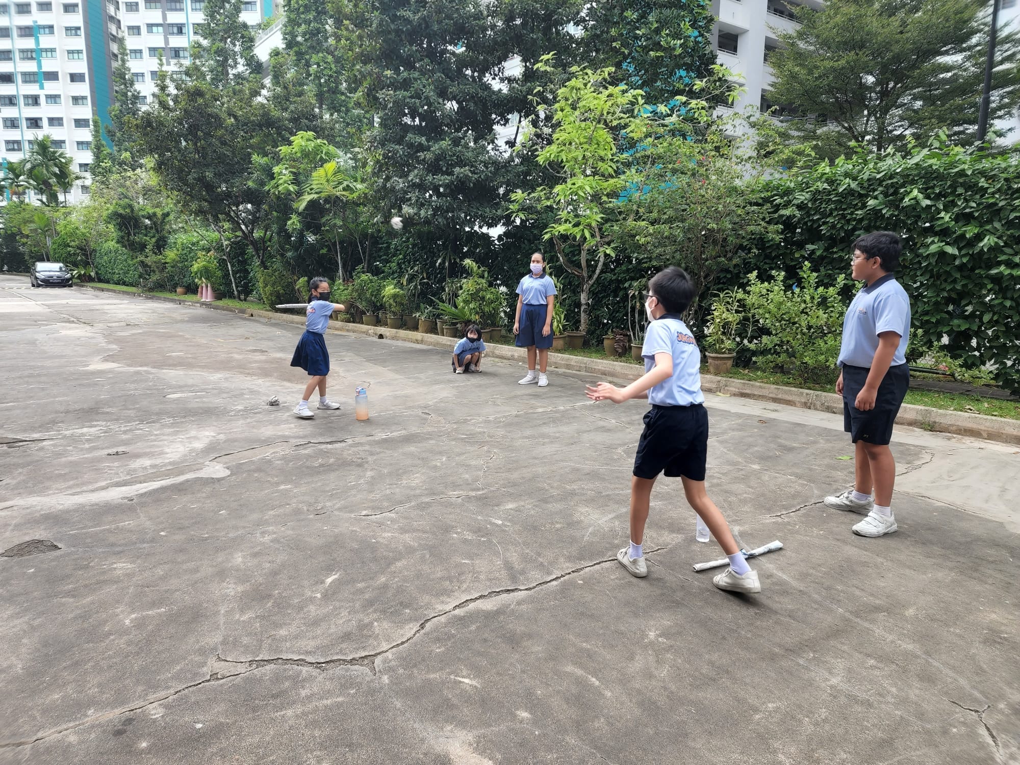 Five children in blue school uniforms play with a ball on an outdoor concrete court surrounded by trees and apartment buildings.