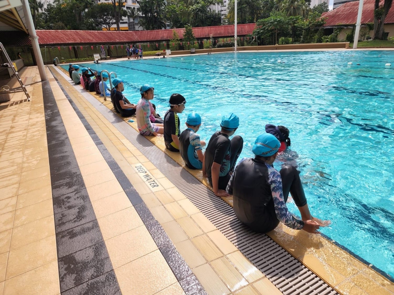 Children in blue swim caps sit along the edge of an outdoor pool labeled "Deep Water," dangling feet in.