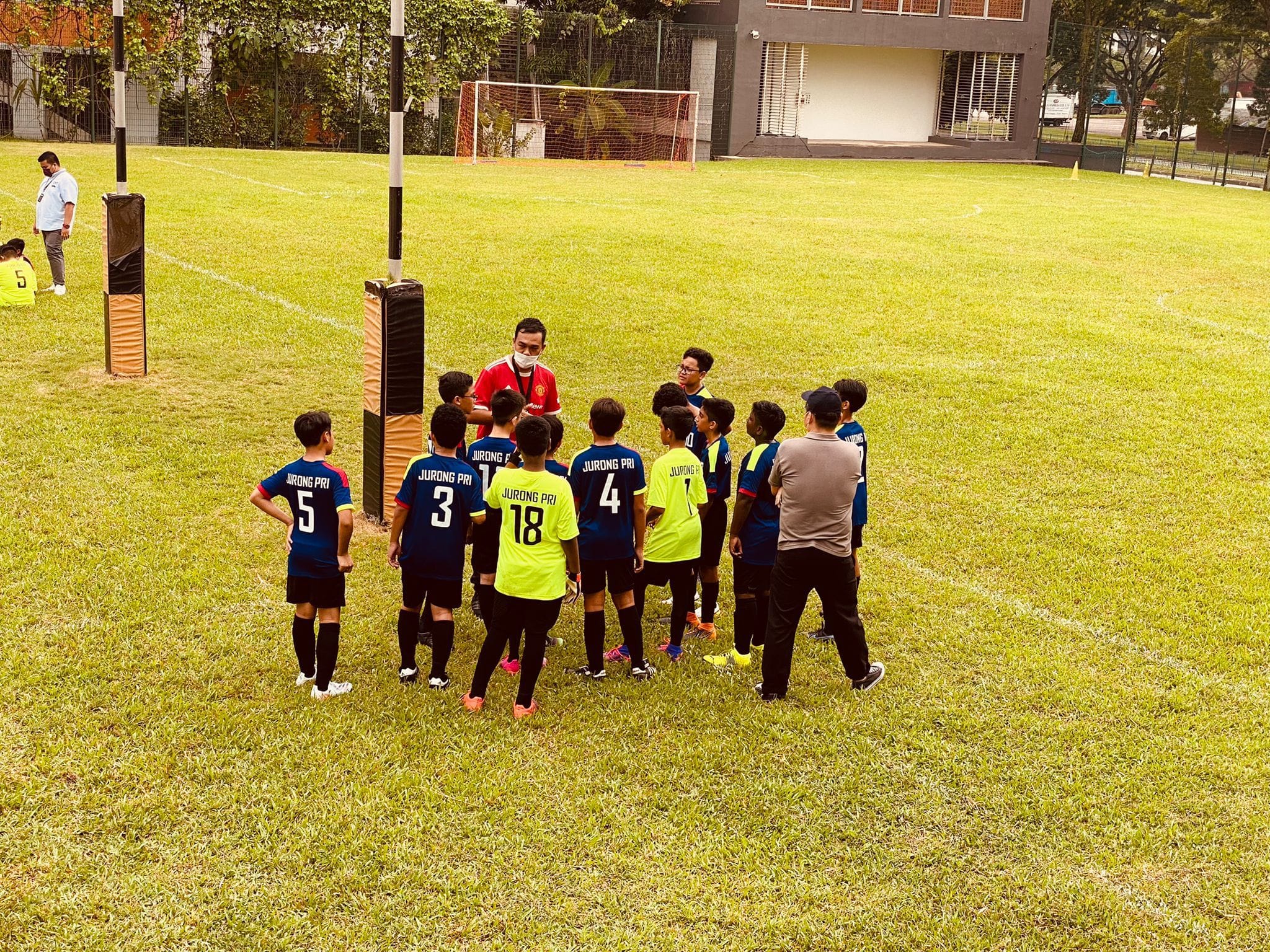 A coach addresses young Jurong Pri football players in blue and yellow jerseys huddled on a green field.