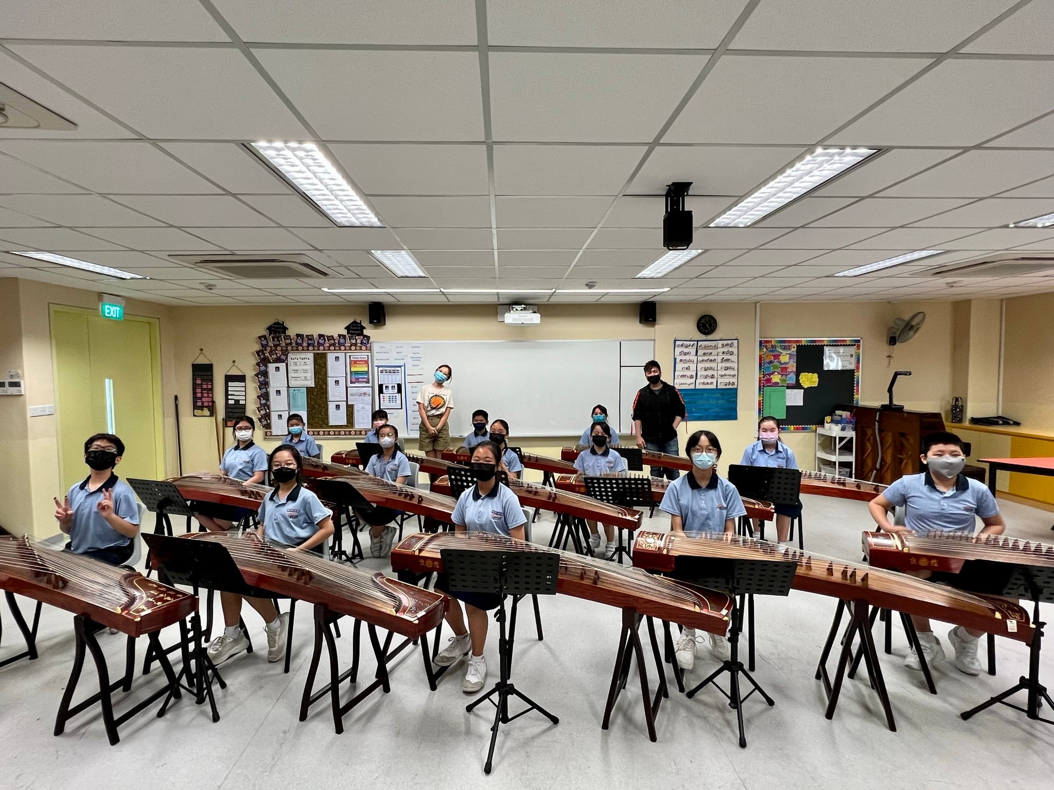 Students in a classroom wearing masks sit at guzheng instruments arranged in rows, posing for a group photo.