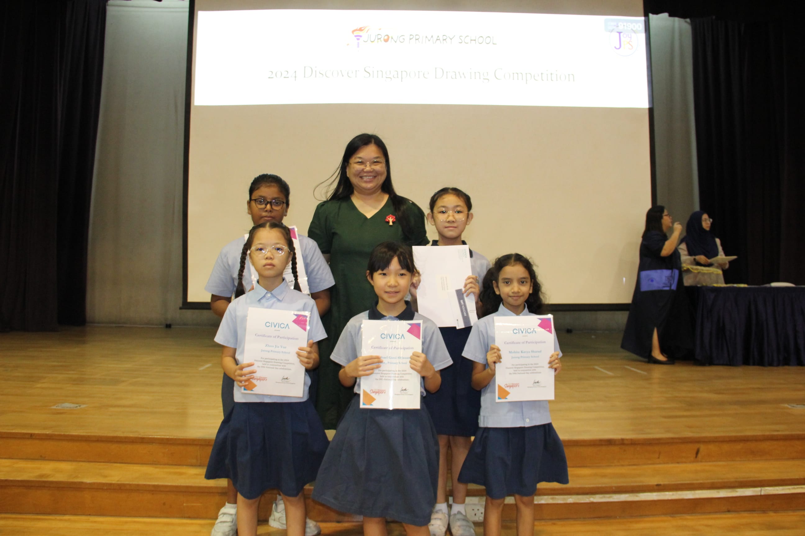 A colour photograph of five young students in light blue school uniforms posing on a school stage with a female teacher in a dark green dress. Three students in the front row hold CIVICA certificates of participation. A projection screen behind them reads "Jurong Primary School — 2024 Discover Singapore Drawing Competition." The group is celebrating their participation in the drawing competition.