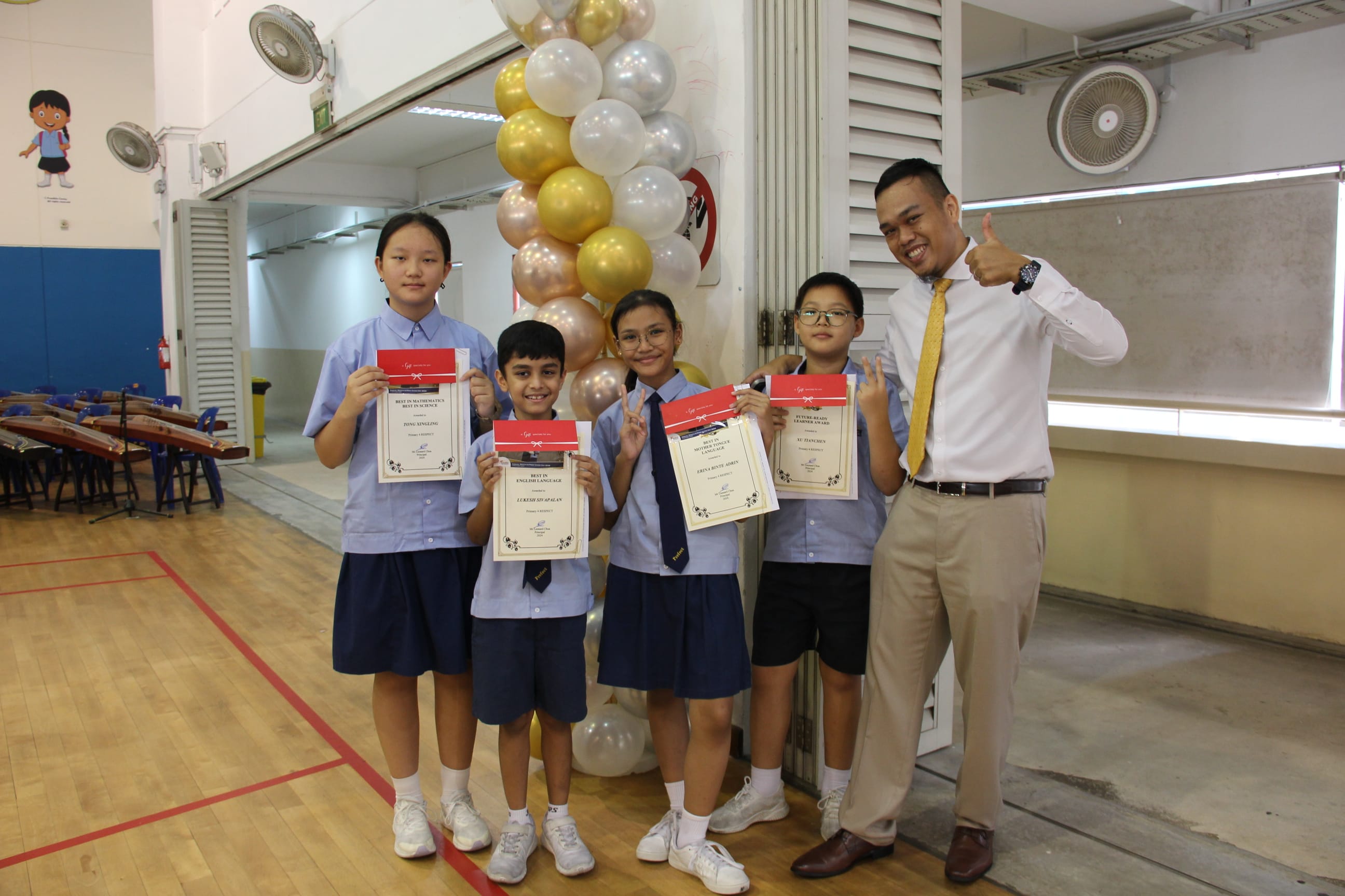 A colour photograph of four students in blue school uniforms and a male teacher in a white shirt and gold tie, posing together in a school hall. Each student holds a certificate with a red ribbon. The teacher stands to the right, smiling and giving a thumbs-up. A tall balloon column in gold, silver, and white decorates the background. Musical instruments are visible on the left.