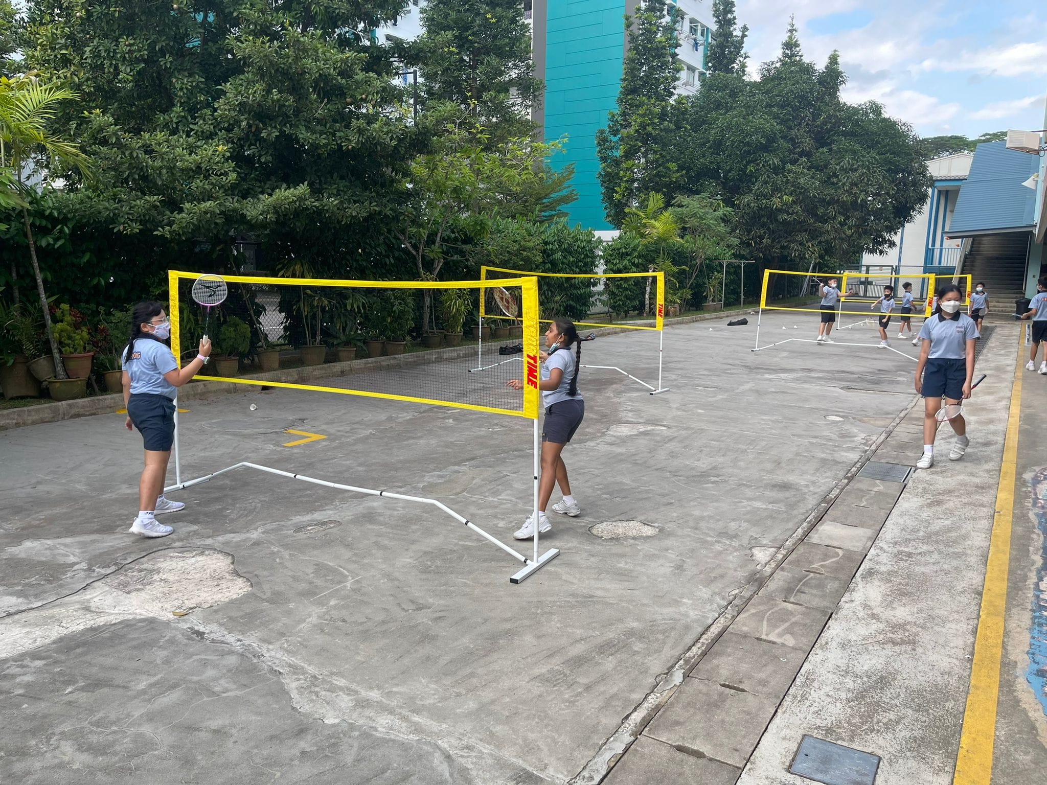 Students in uniforms and masks play badminton across yellow portable nets on an outdoor concrete court.