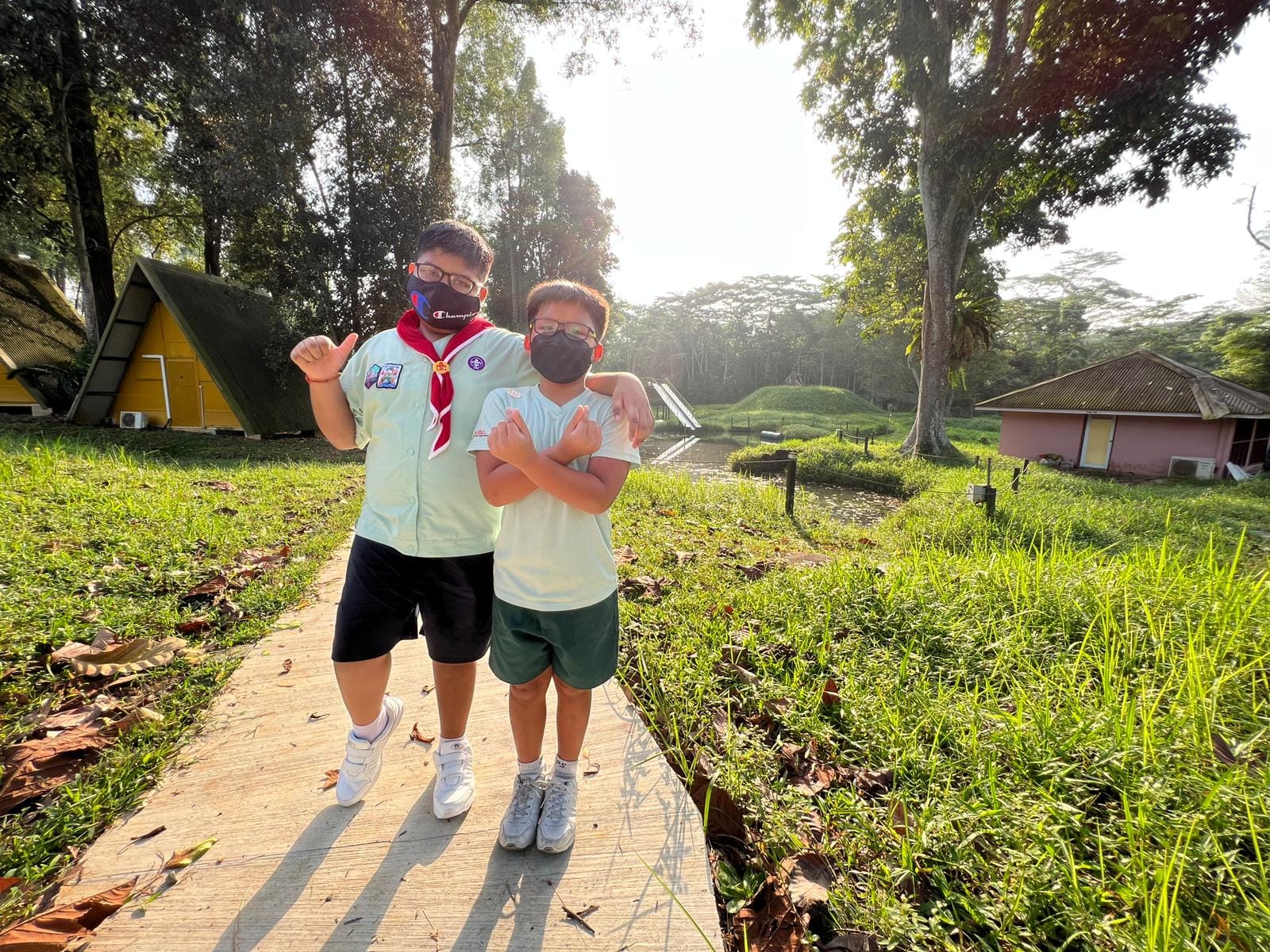 Two masked boys in scout uniforms pose on a sunlit path surrounded by green grass, trees, and small cabin buildings.