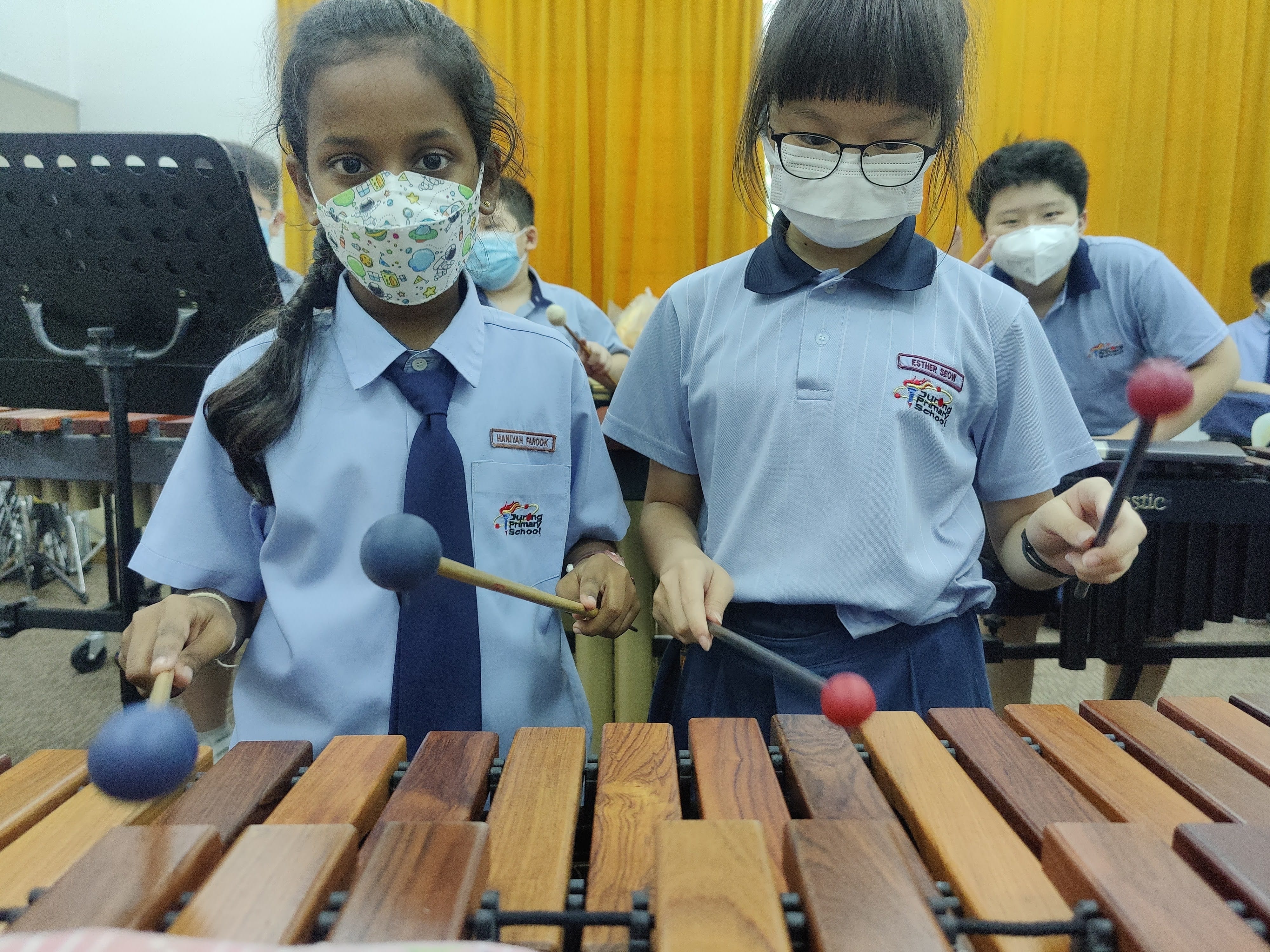 Two masked primary school girls in blue uniforms play a marimba with mallets in a classroom setting.