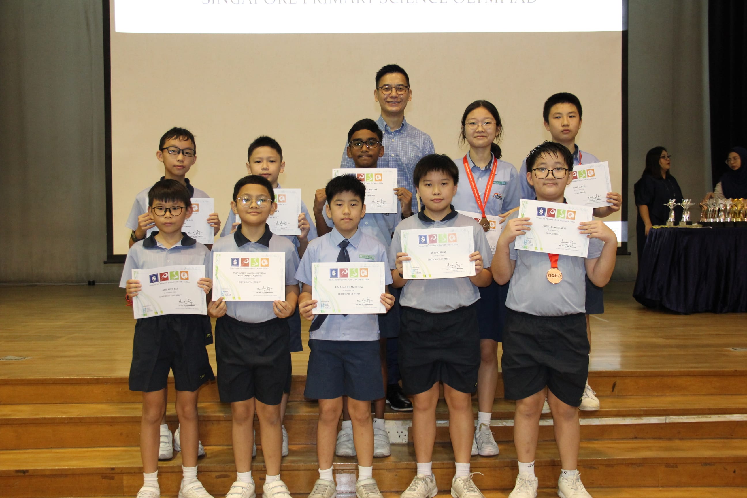 A colour photograph of nine students in light blue school uniforms posing on a school stage with a male teacher. Each student holds a certificate from the Singapore Primary Science Olympiad, as indicated by the partially visible projection screen above. Two students wear bronze medals around their necks. Trophies are visible on a table to the right. The group is celebrating their achievement in the science competition.