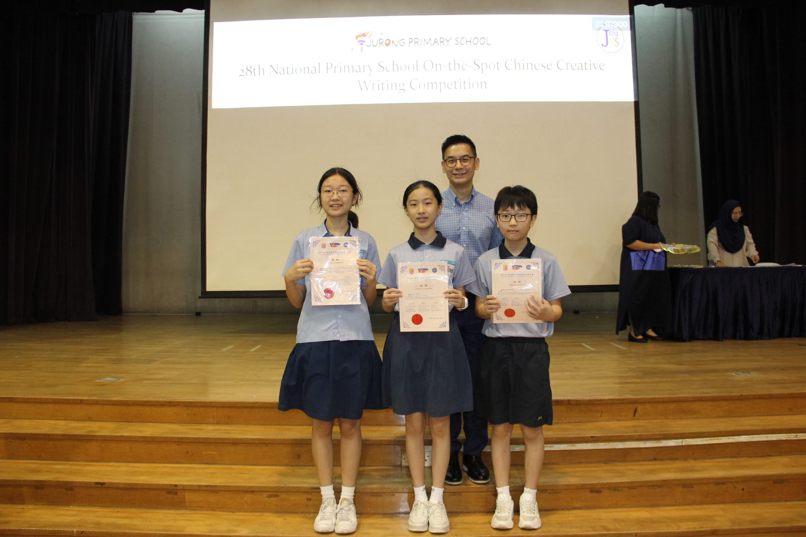 A colour photograph of three students in light blue school uniforms posing on a school stage with a male teacher. Each student holds a certificate with a red seal. A projection screen behind them reads "Jurong Primary School — 28th National Primary School On-the-Spot Chinese Creative Writing Competition." The group is celebrating their achievement in the Chinese creative writing competition.
