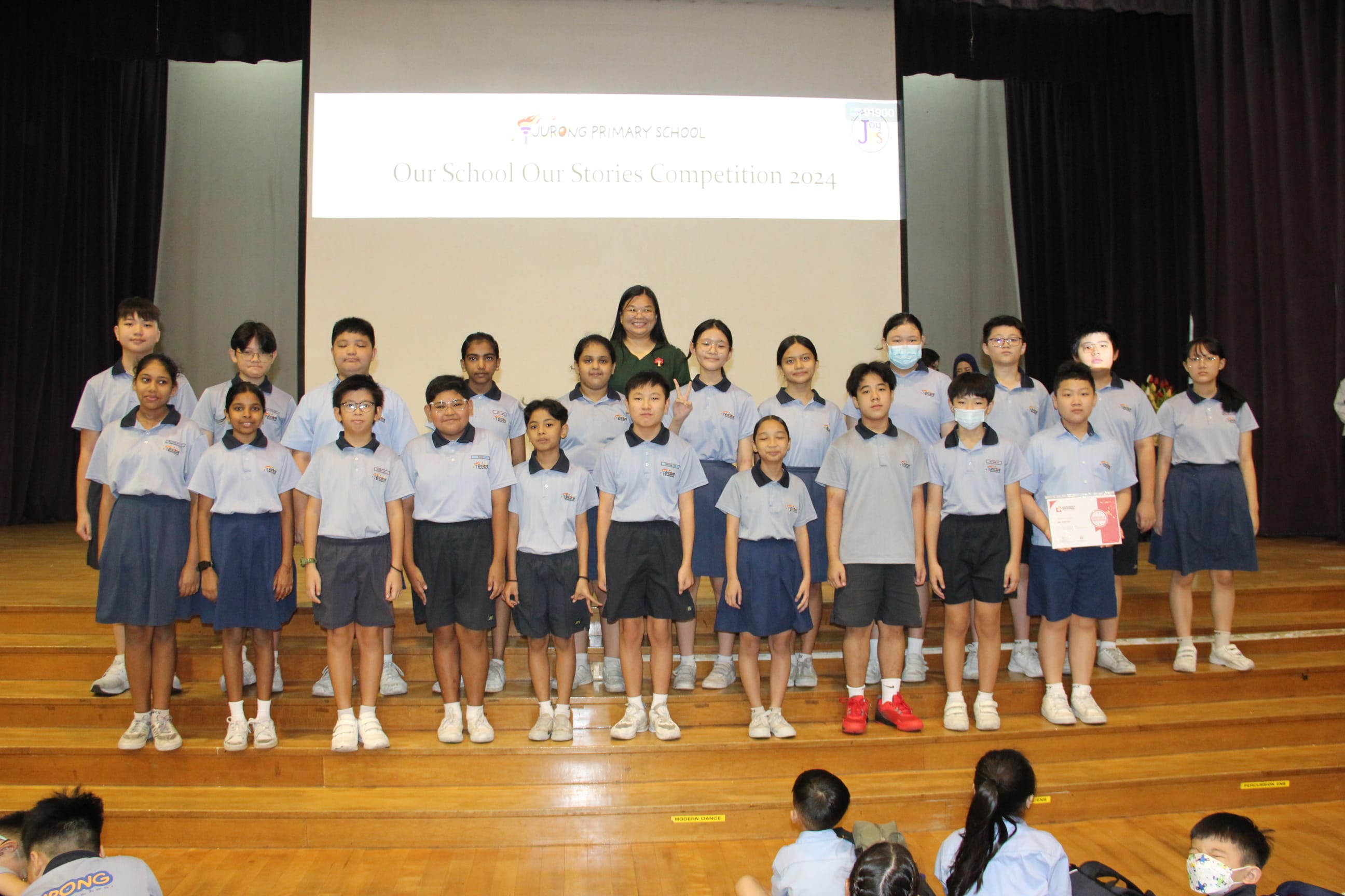 A colour photograph of approximately twenty-five students in light blue school uniforms posing on a school stage with a female teacher in a dark green dress. One student holds a certificate. A projection screen behind them reads "Jurong Primary School — Our School Our Stories Competition 2024." The diverse group of students, ranging from younger to older primary school age, is celebrating their participation in the storytelling competition.