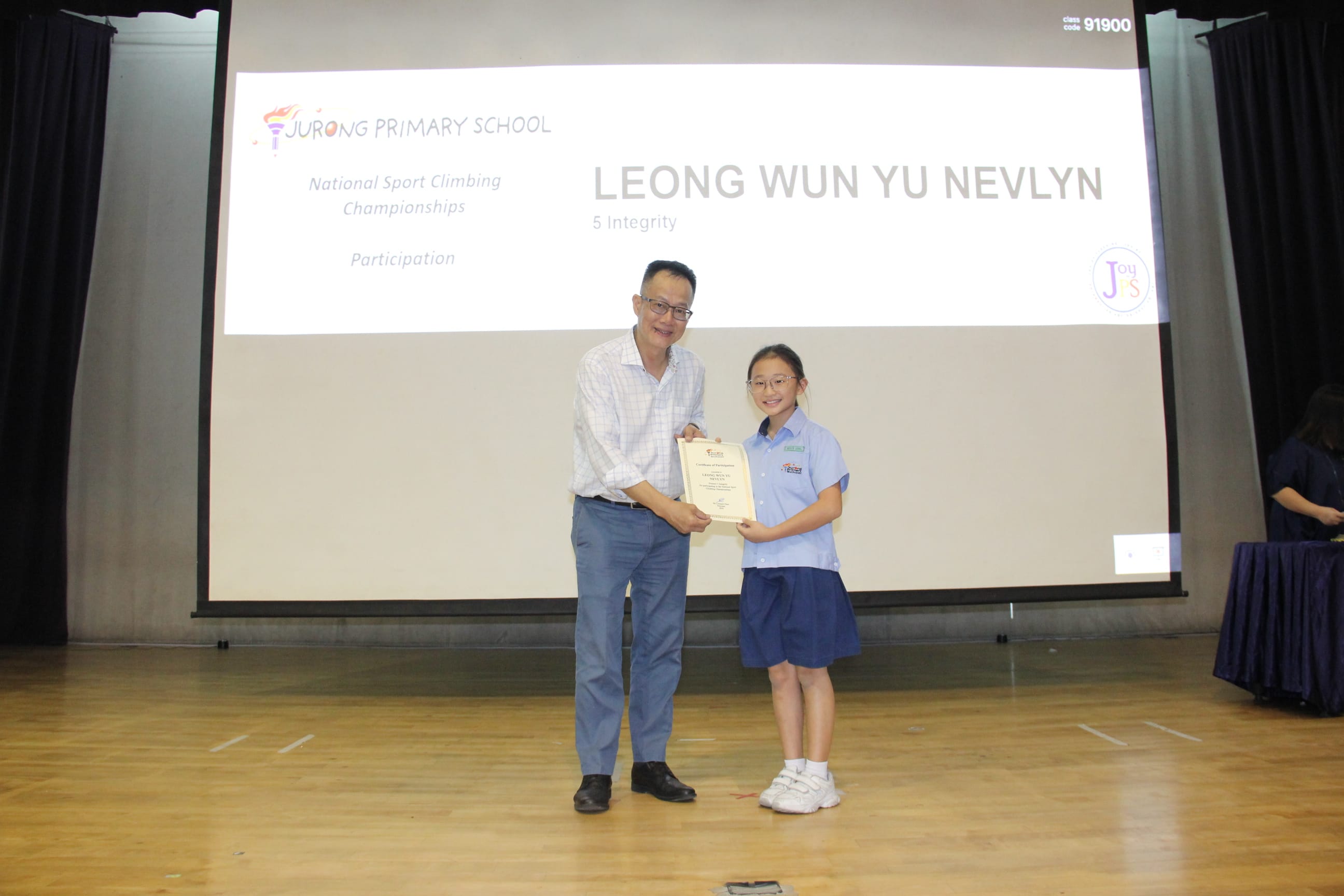 A colour photograph taken on a school stage showing a smiling male teacher handing a participation certificate to a young female student in a light blue school uniform. Behind them, a projection screen displays "Jurong Primary School — National Sport Climbing Championships, Participation" alongside the student's name "Leong Wun Yu Nevlyn, 5 Integrity," honouring her participation in the sport climbing championships.
