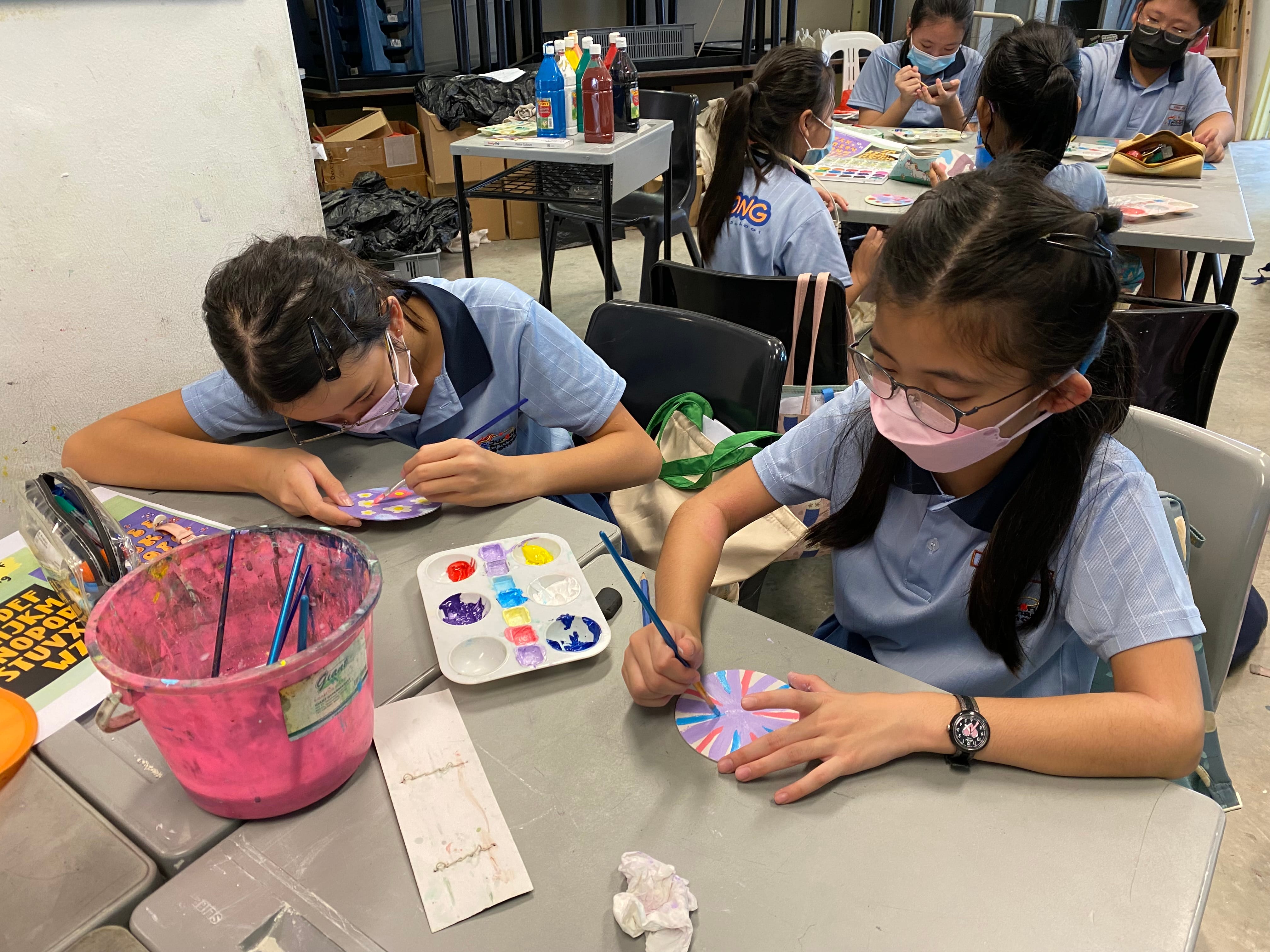 Students in school uniforms and masks paint colorful circular designs at tables with palettes and brushes.