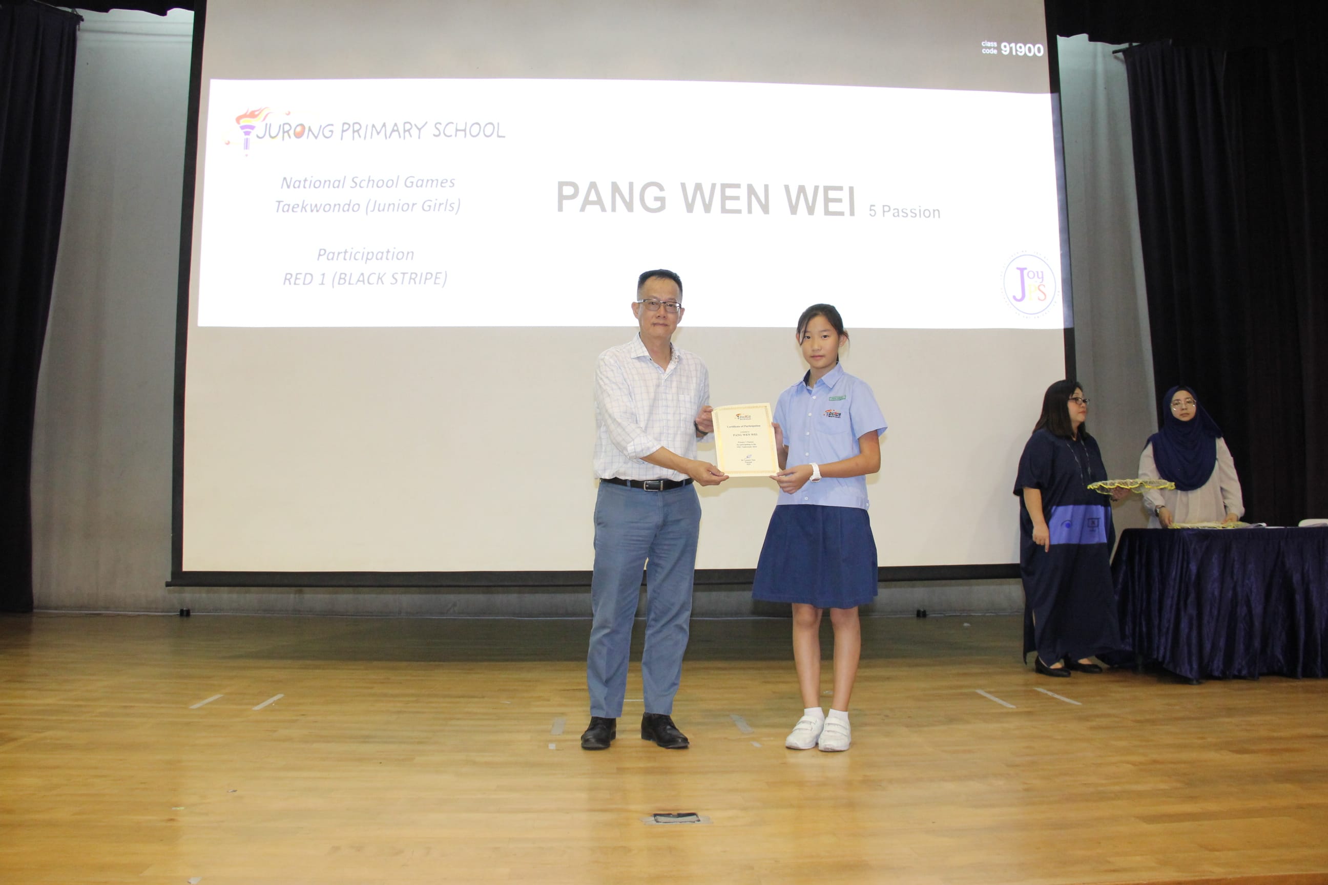A colour photograph of a male teacher in a light checked shirt presenting a certificate to a female student in a light blue school uniform on a school stage. A projection screen behind them reads "Jurong Primary School — National School Games Taekwondo (Junior Girls), Participation, RED 1 (BLACK STRIPE)" with the student's name "Pang Wen Wei, 5 Passion" displayed. The student is being recognised for her participation in the taekwondo competition.