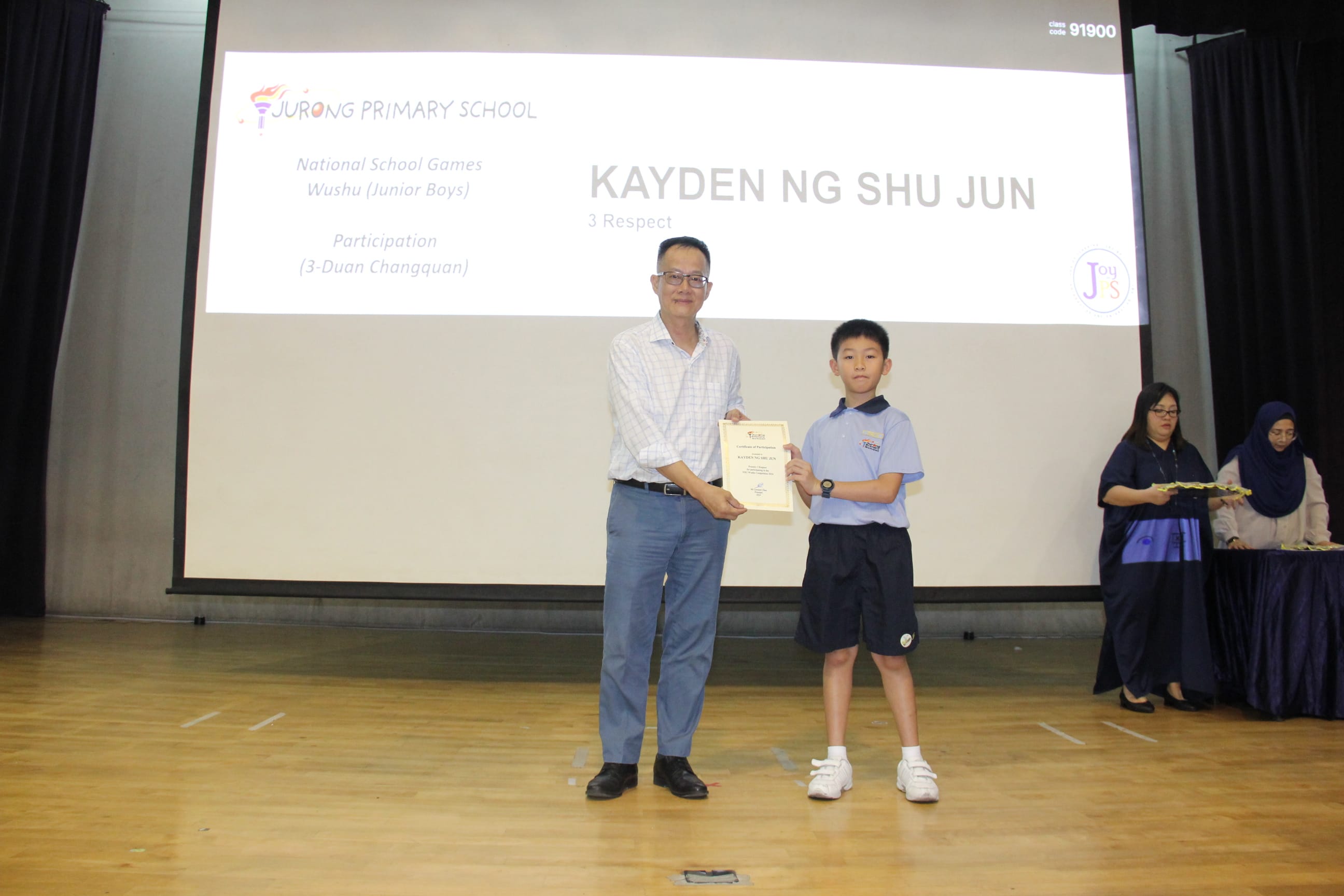 It looks like this is the same image as the previous one! Here is the same description:  A colour photograph of a male teacher in a light checked shirt presenting a certificate to a young male student in a light blue school uniform on a school stage. A projection screen behind them reads "Jurong Primary School — National School Games Wushu (Junior Boys), Participation (3-Duan Changquan)" with the student's name "Kayden Ng Shu Jun, 3 Respect" displayed. The student is being recognised for his participation in the wushu competition.