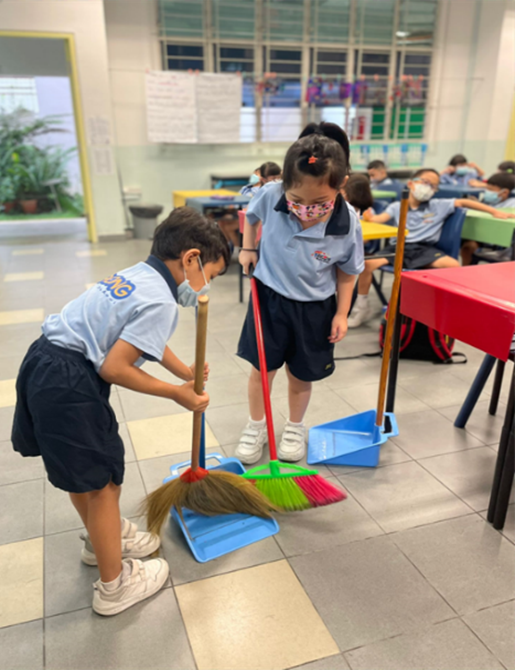 Two young students in school uniforms and masks sweep the classroom floor with brooms and dustpans.
