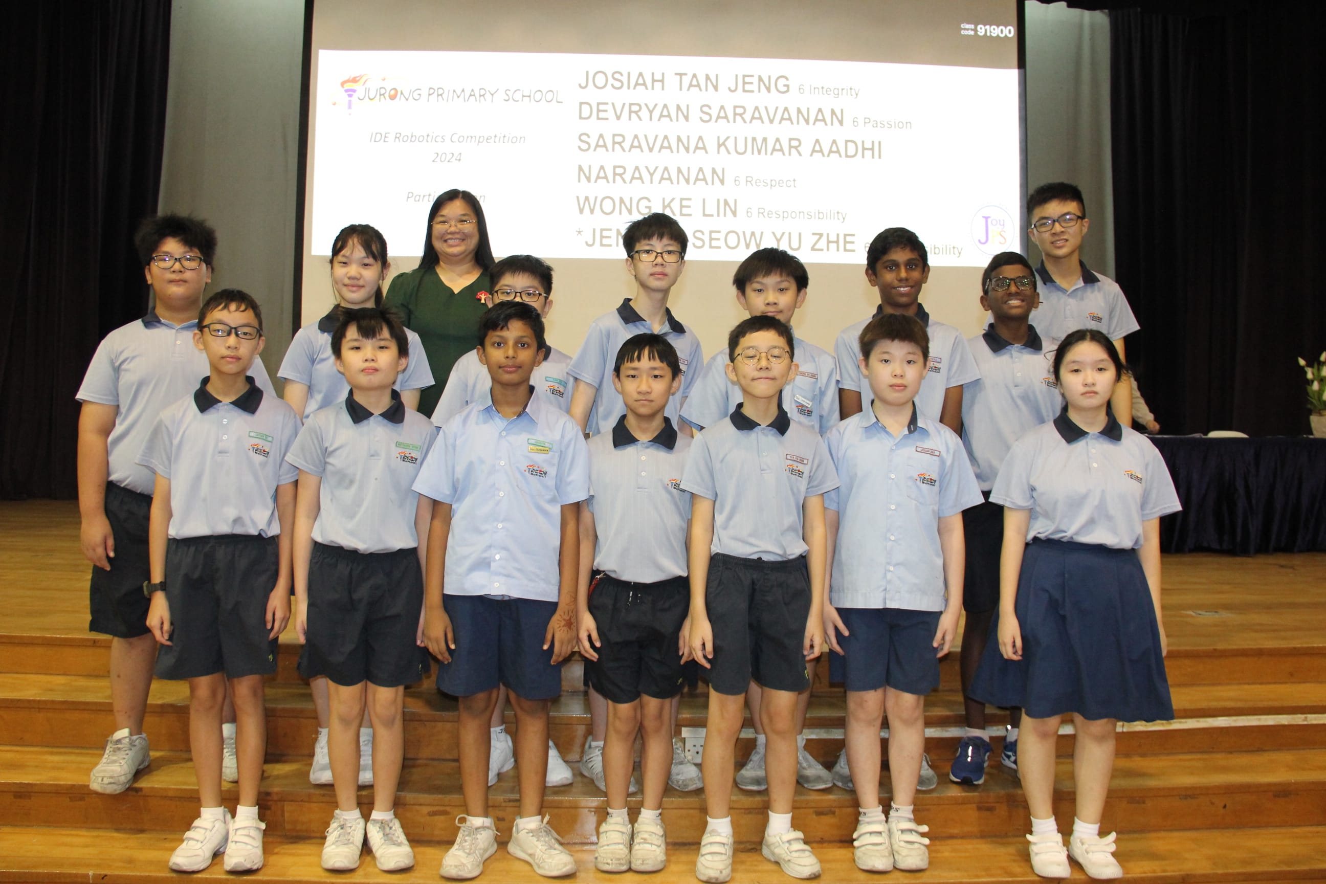 A colour photograph of approximately fifteen students in light blue school uniforms posing on a school stage with a female teacher in a dark green dress. A projection screen behind them reads "Jurong Primary School — IDE Robotics Competition 2024," with a list of participating students' names and classes visible. The diverse group of students, ranging from younger to older primary school age, is celebrating their participation in the robotics competition.