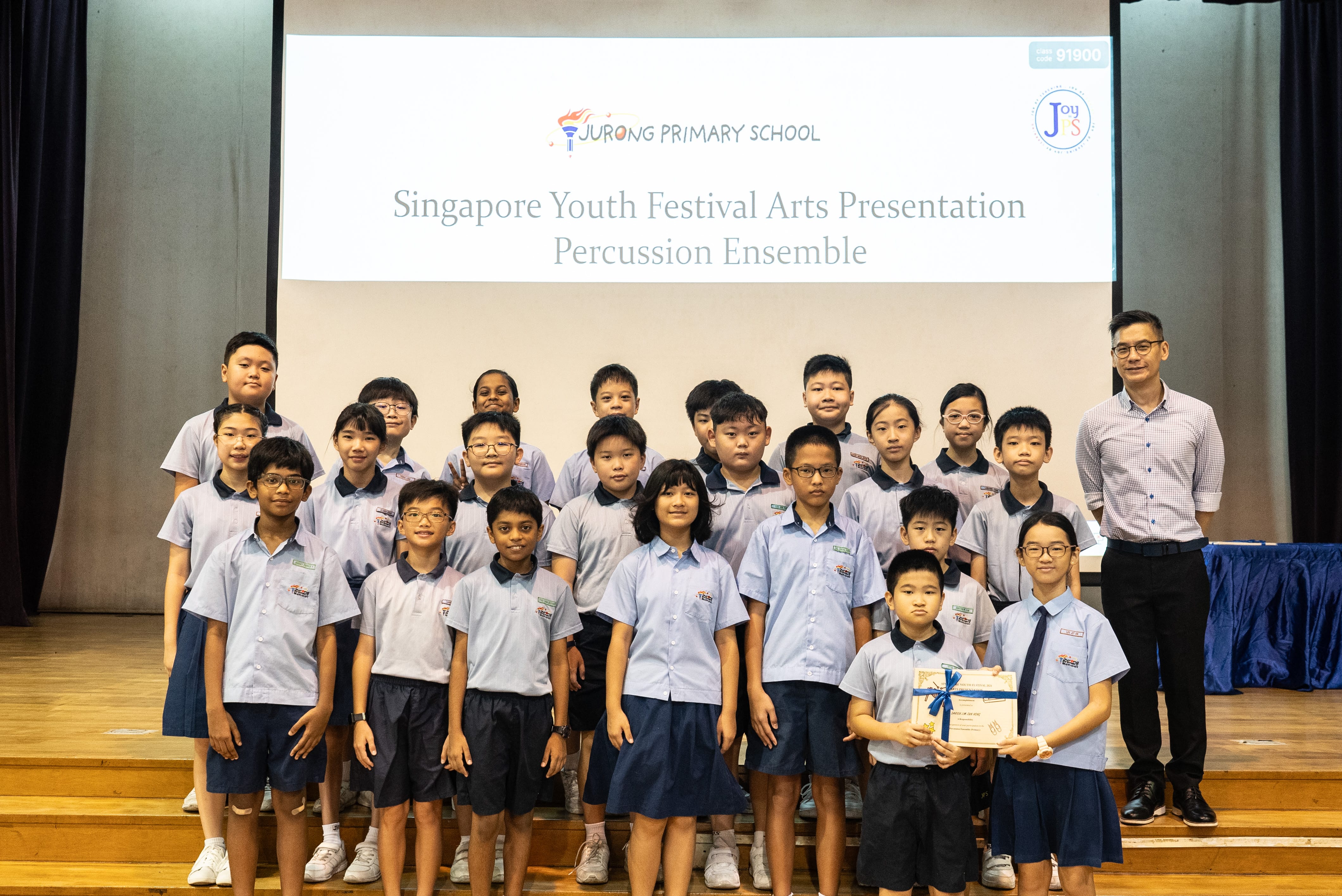 A colour photograph of a group of approximately twenty students in light blue school uniforms posing on a school stage with a male teacher. A projection screen behind them reads "Jurong Primary School — Singapore Youth Festival Arts Presentation, Percussion Ensemble." One student in the front row holds a certificate tied with a blue ribbon. The group appears to be celebrating their participation or achievement in the arts event.