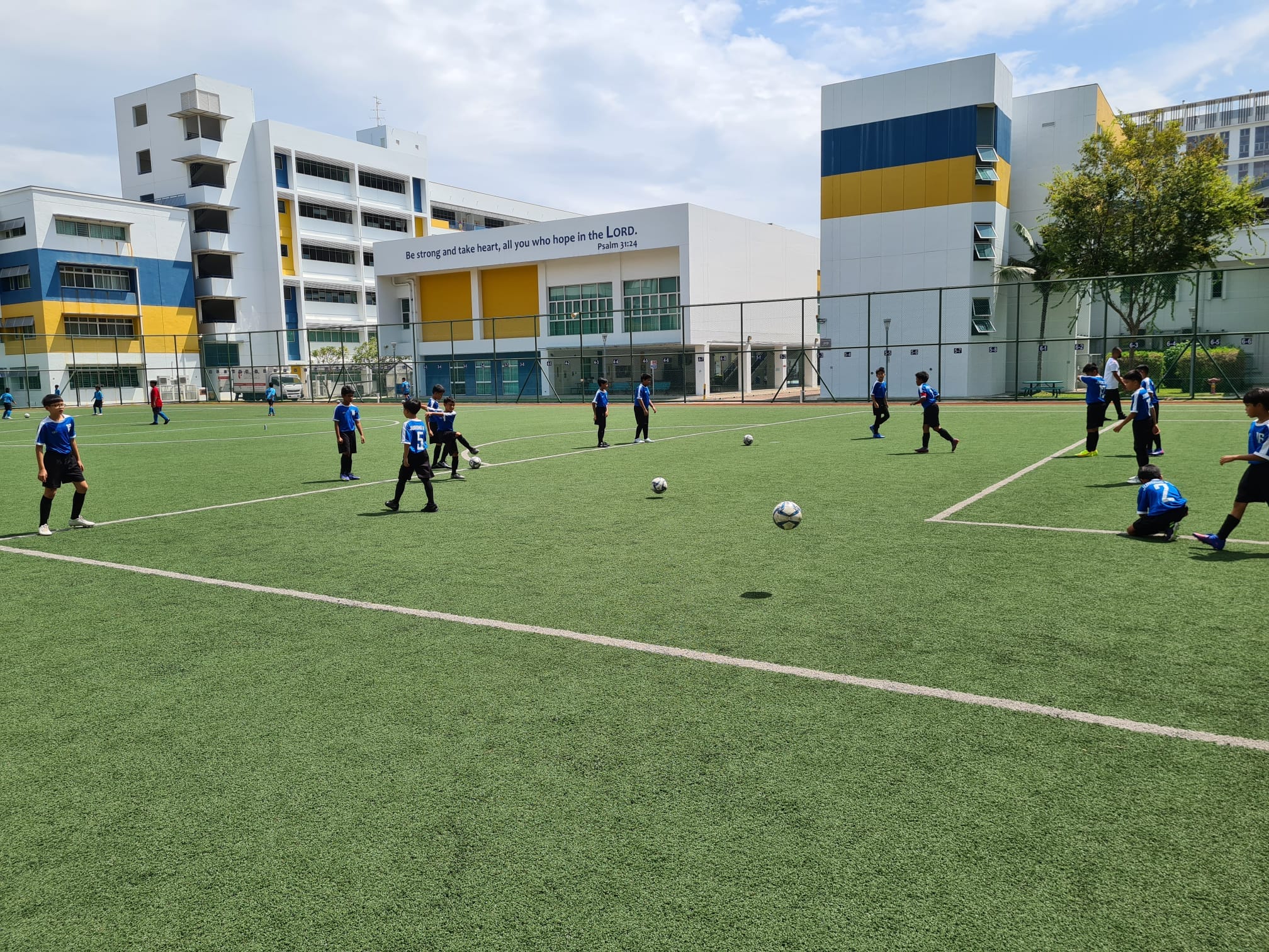Boys in blue jerseys play soccer on a green artificial turf field beside multi-story school buildings.
