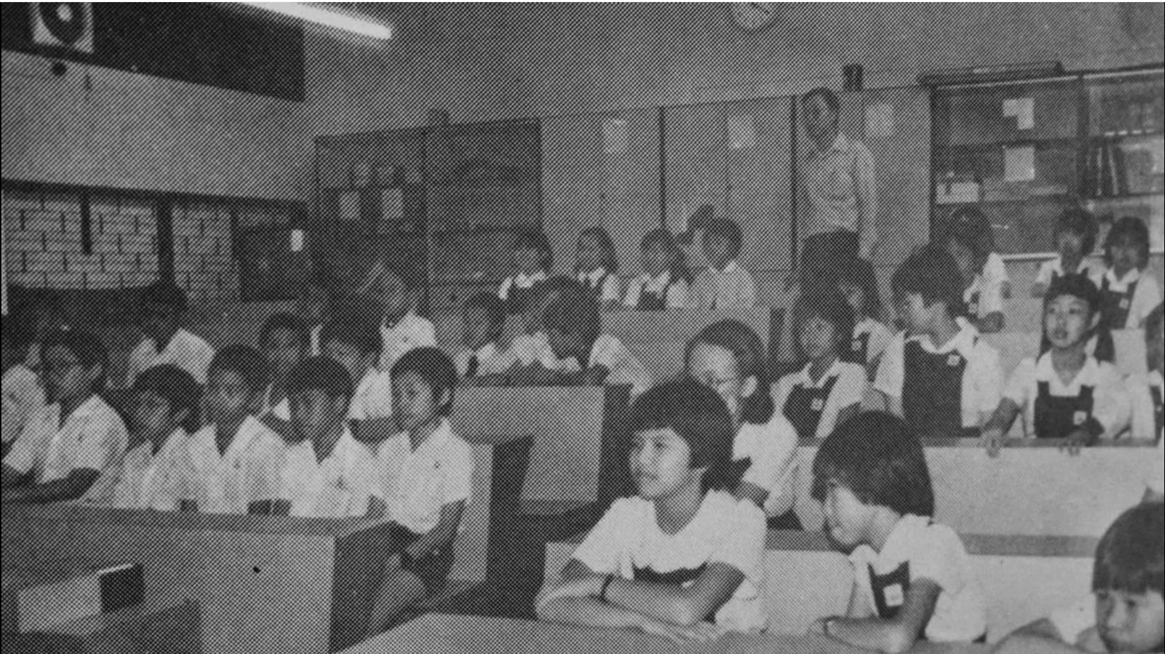 A black-and-white vintage photograph of a large classroom filled with young students in school uniforms — white shirts and dark pinafores — seated at rows of desks. The children appear attentive, facing towards the front of the room. A television set is mounted in the upper left corner, suggesting a lesson being conducted via broadcast. The room is well-lit with fluorescent lighting, and louvred windows line the walls in the background. The setting appears to be a primary school from several decades ago, likely in Southeast Asia.