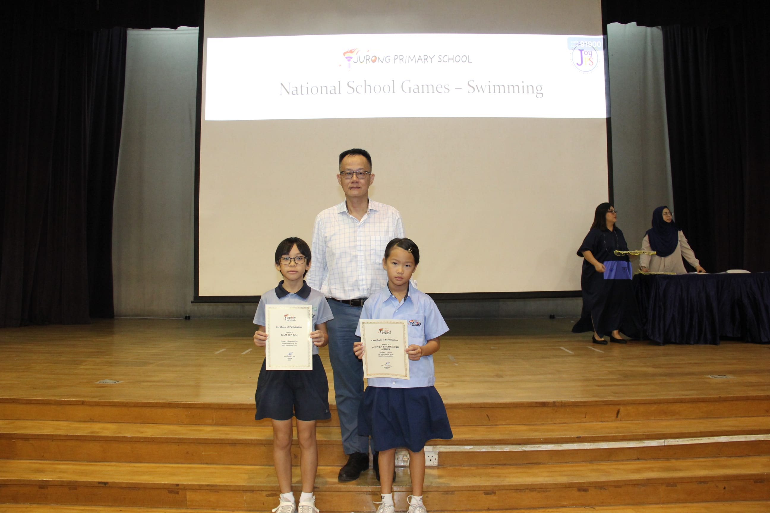 A colour photograph of two young female students in light blue school uniforms posing on a school stage with a male teacher in a light checked shirt. Each student holds a certificate of participation. A projection screen behind them reads "Jurong Primary School — National School Games – Swimming." The group is celebrating the students' participation in the swimming competition.