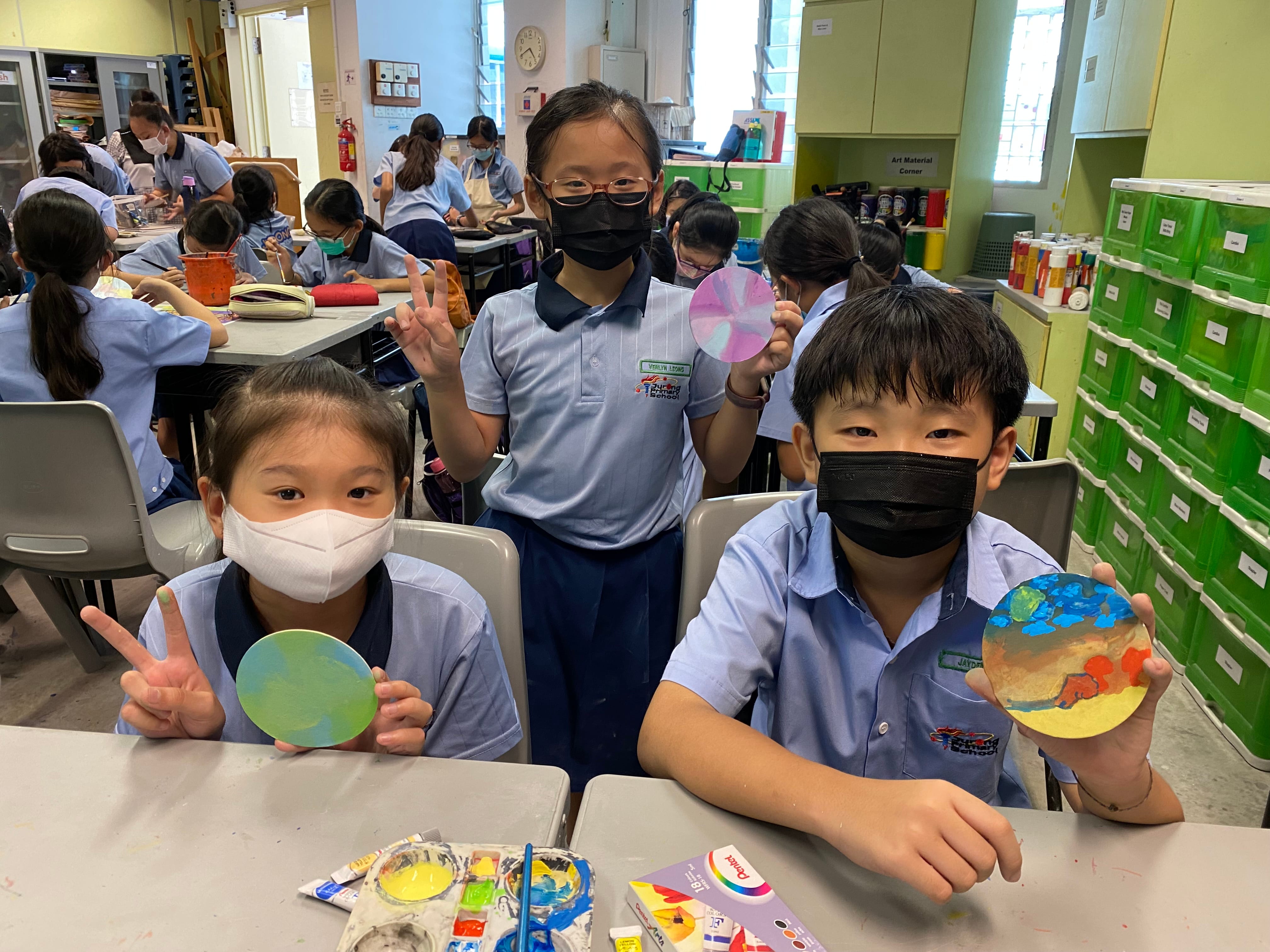 Three masked school children hold up colorful painted circular artworks in a busy art classroom.