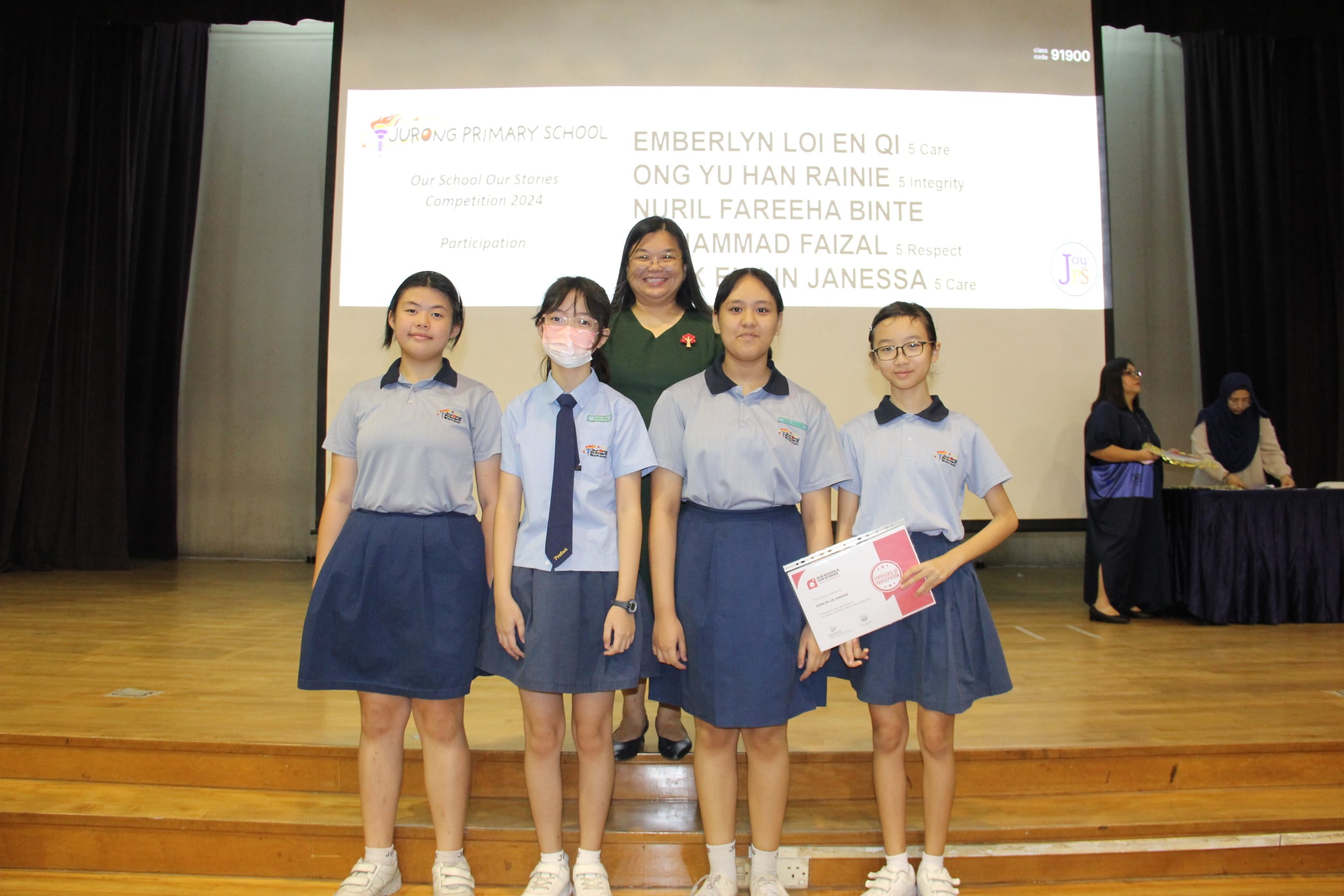 A colour photograph of four female students in light blue school uniforms posing on a school stage with a female teacher in a dark green dress. One student holds a certificate. A projection screen behind them reads "Jurong Primary School — Our School Our Stories Competition 2024, Participation," with the students' names listed. The group is celebrating their participation in the storytelling competition.