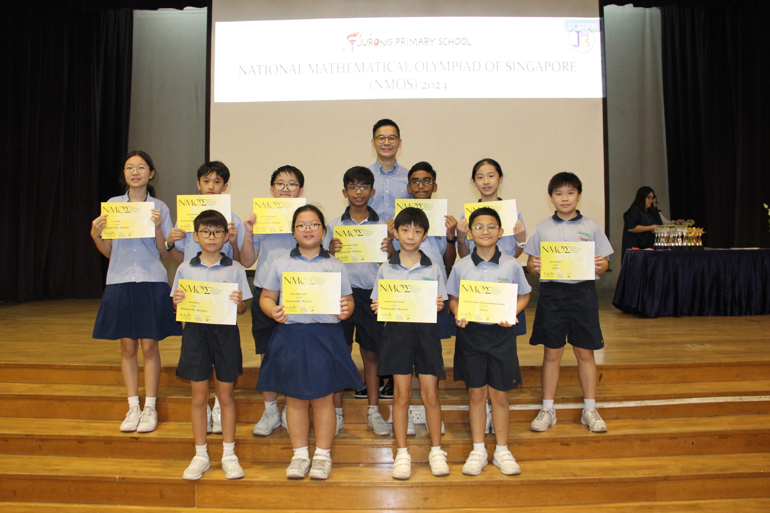 A colour photograph of eleven students in light blue school uniforms posing on a school stage with a male teacher. Each student proudly holds a yellow NMOS certificate. A projection screen behind them reads "Jurong Primary School — National Mathematical Olympiad of Singapore (NMOS) 2024." Trophies are visible on a table to the right. The group is celebrating their achievement in the mathematics competition.