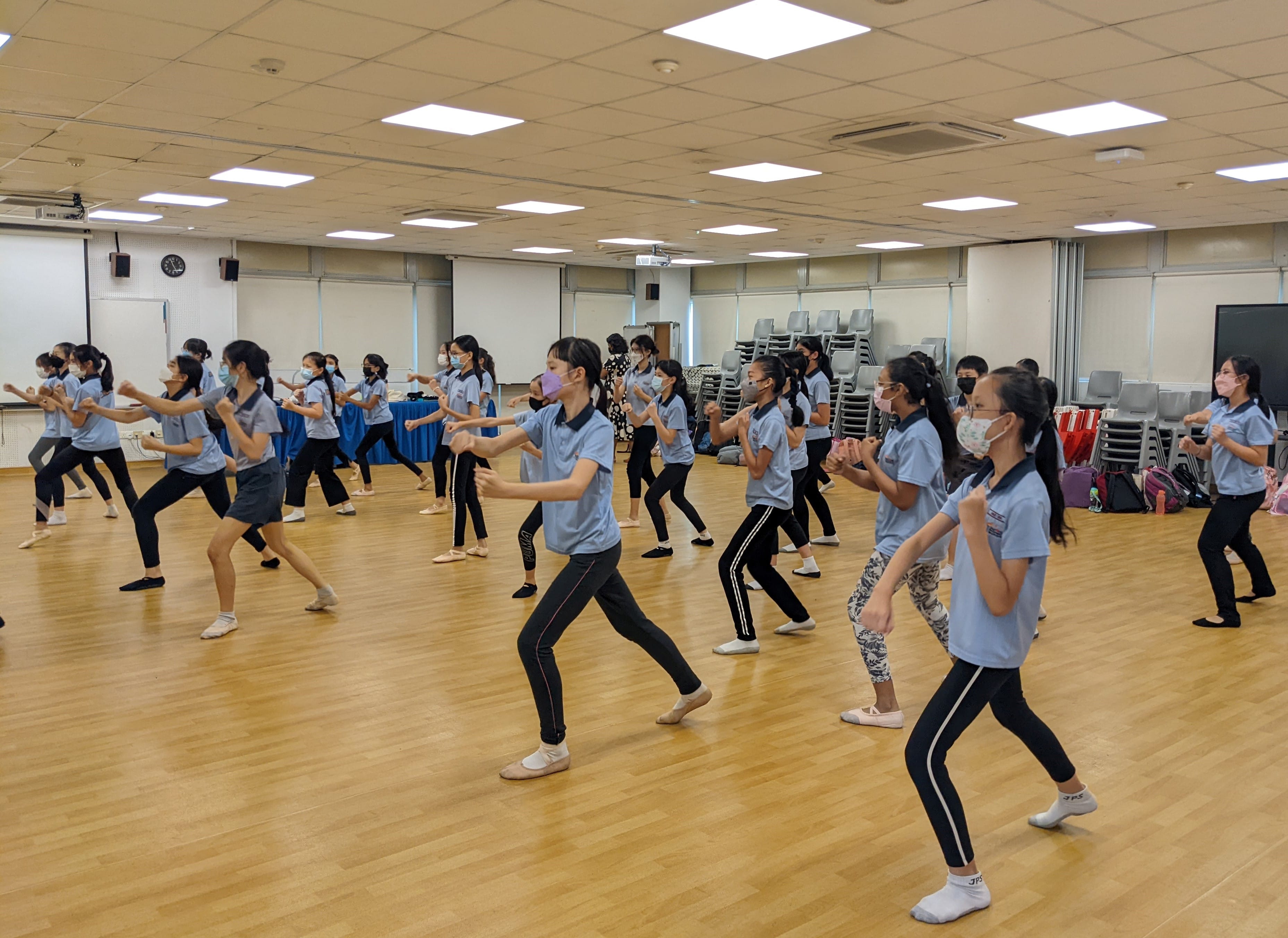 A large group of masked students in light blue shirts practice a dance routine on a wooden floor indoors.