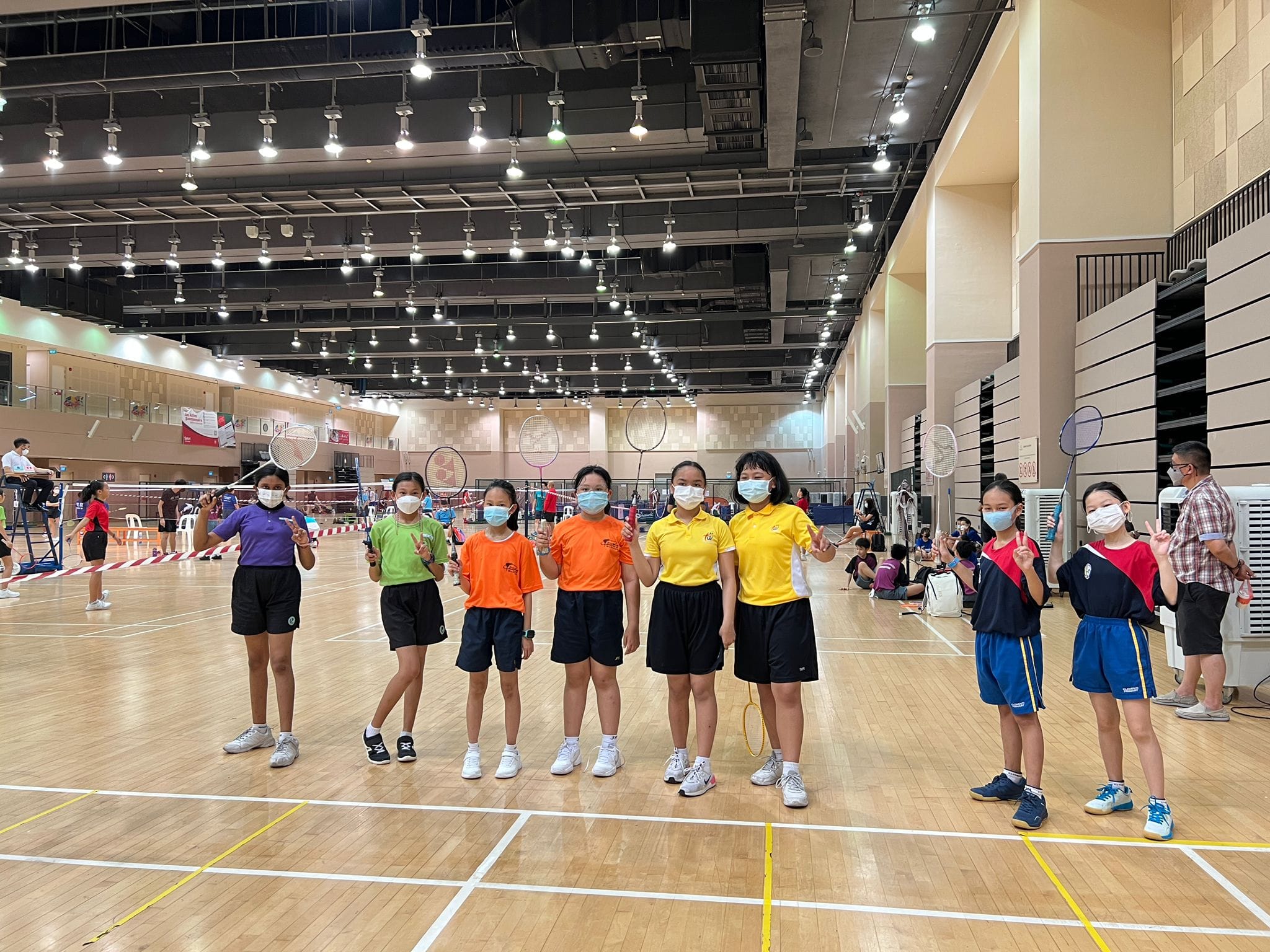 Seven masked young girls holding badminton rackets pose together on an indoor badminton court.