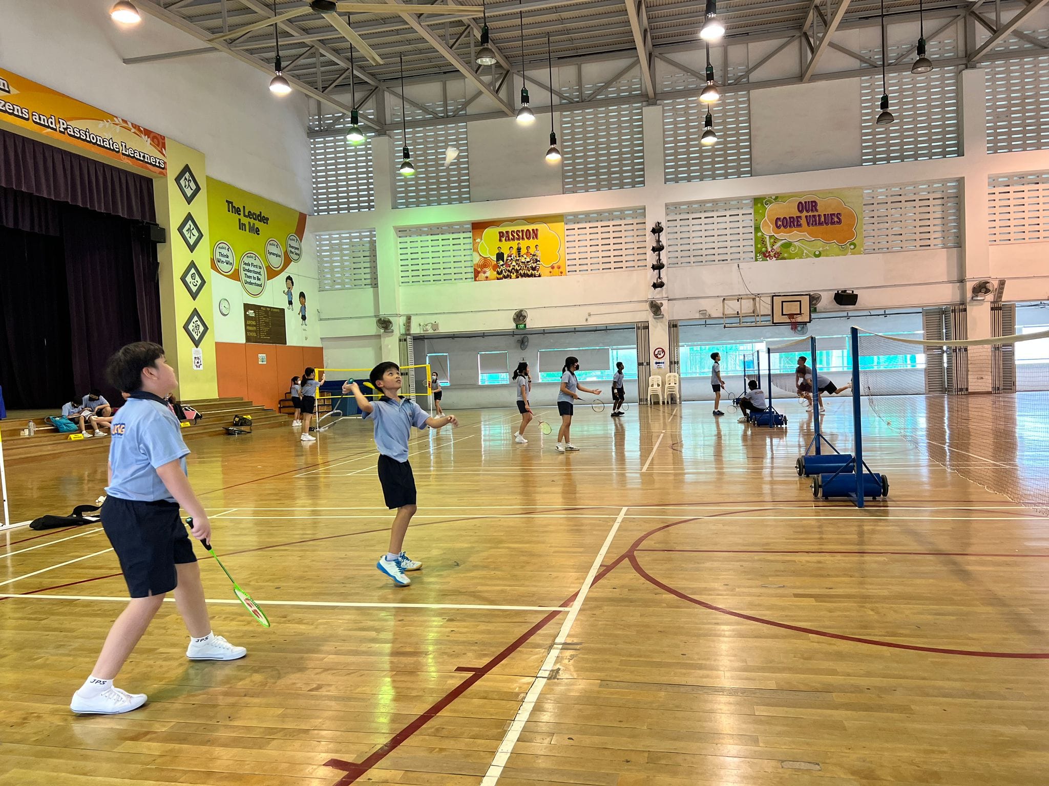 Children in blue uniforms play badminton in a school gymnasium with colorful banners on the walls.