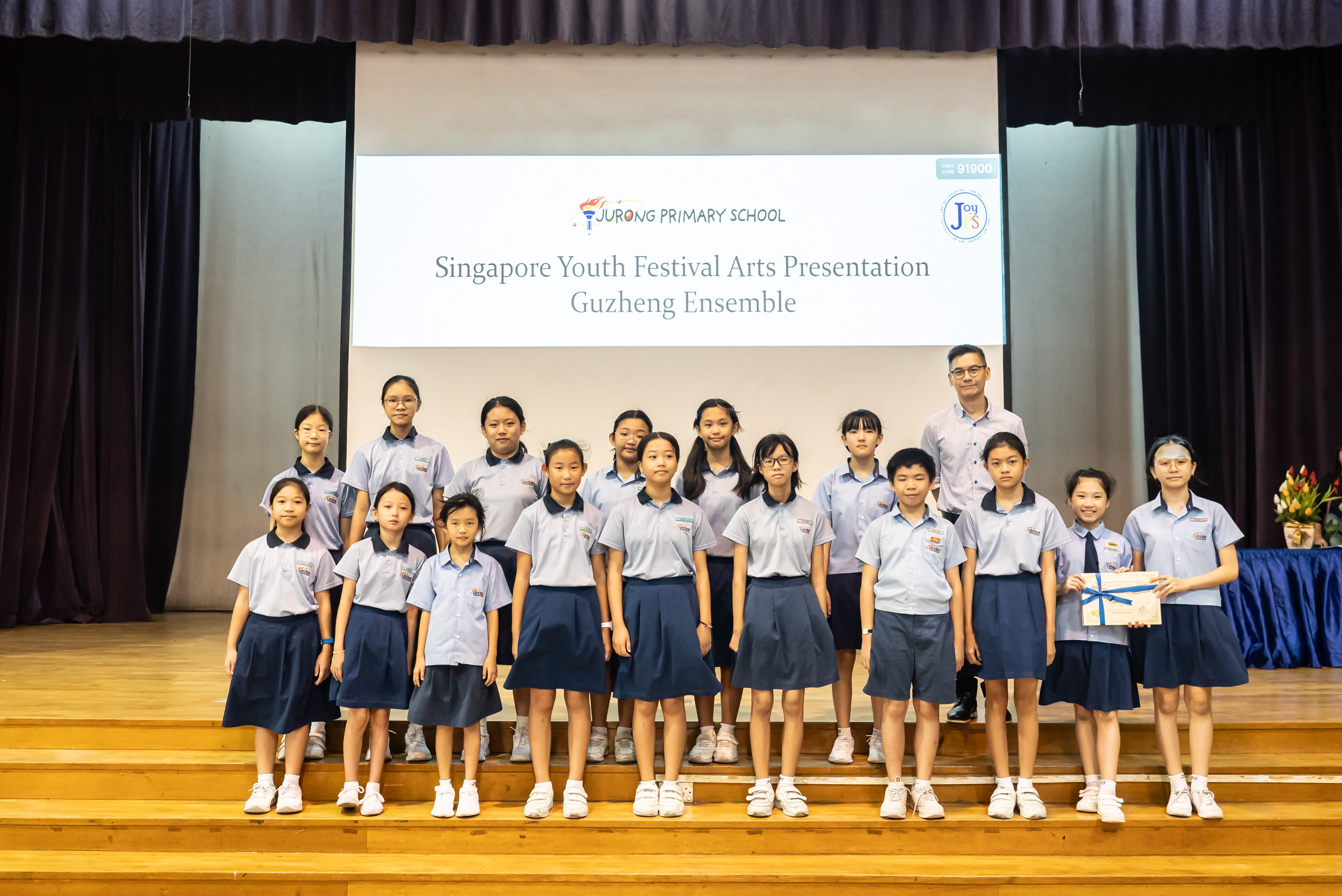 A colour photograph of approximately fifteen students in light blue school uniforms posing on a school stage with a male teacher. A projection screen behind them reads "Jurong Primary School — Singapore Youth Festival Arts Presentation, Guzheng Ensemble." A student on the right holds a certificate tied with a blue ribbon. The group is celebrating their participation or achievement in the Chinese instrument ensemble event.