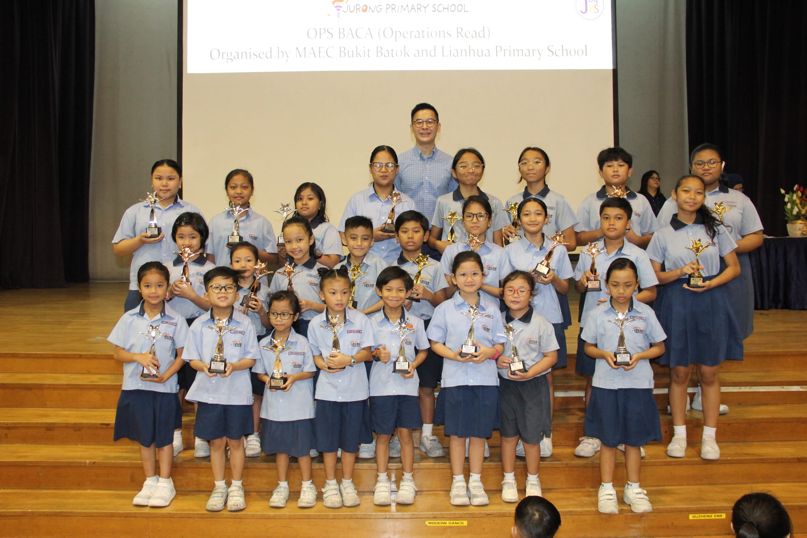 A colour photograph of approximately twenty-five students in light blue school uniforms posing on a school stage with a male teacher. Each student proudly holds a gold star trophy. A projection screen behind them reads "Jurong Primary School — OPS BACA (Operations Read), Organised by MAEC Bukit Batok and Lianhua Primary School." The students range from younger to older primary school age, celebrating their achievement in the Malay language reading competition.
