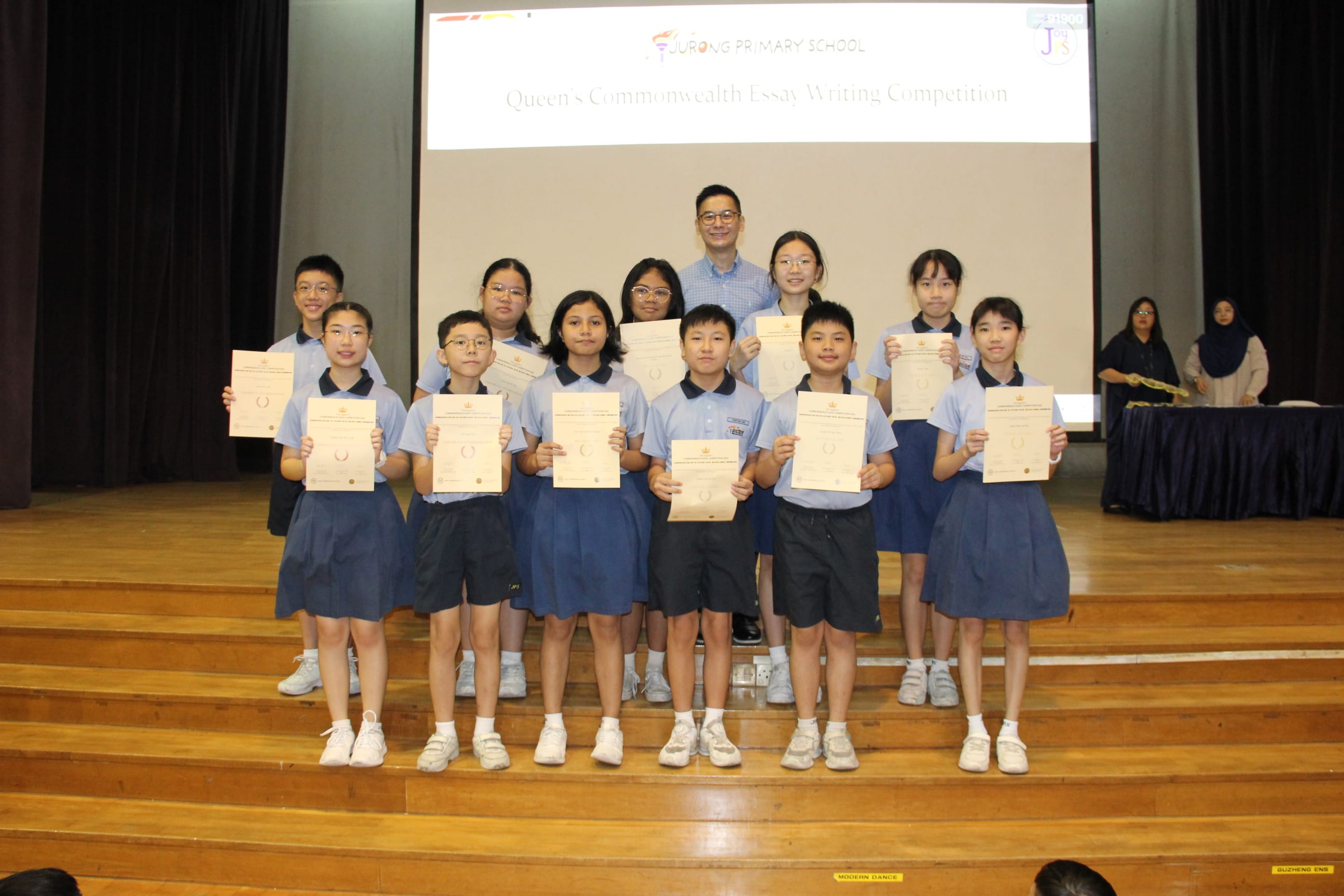 A colour photograph of eleven students in light blue school uniforms posing on a school stage with a male teacher. Each student holds a certificate with a gold emblem. A projection screen behind them reads "Jurong Primary School — Queen's Commonwealth Essay Writing Competition." The group is celebrating their achievement in the English essay writing competition.