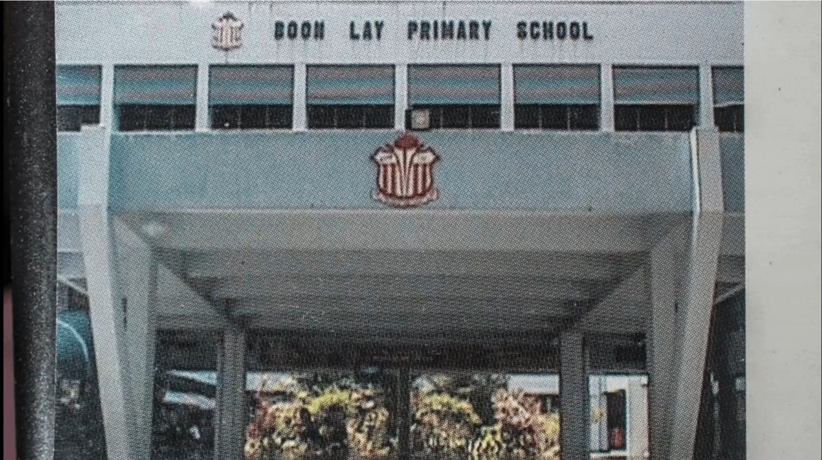 A vintage photograph showing the entrance of Boon Lay Primary School. The school's name is displayed prominently in bold lettering across the top of the building's facade, with the school crest — a red and white shield emblem — mounted below it. The entrance features a covered porch supported by concrete columns, with glass doors through which tropical plants and foliage are visible. The architectural style is typical of Singapore school buildings from the 1970s to 1980s.