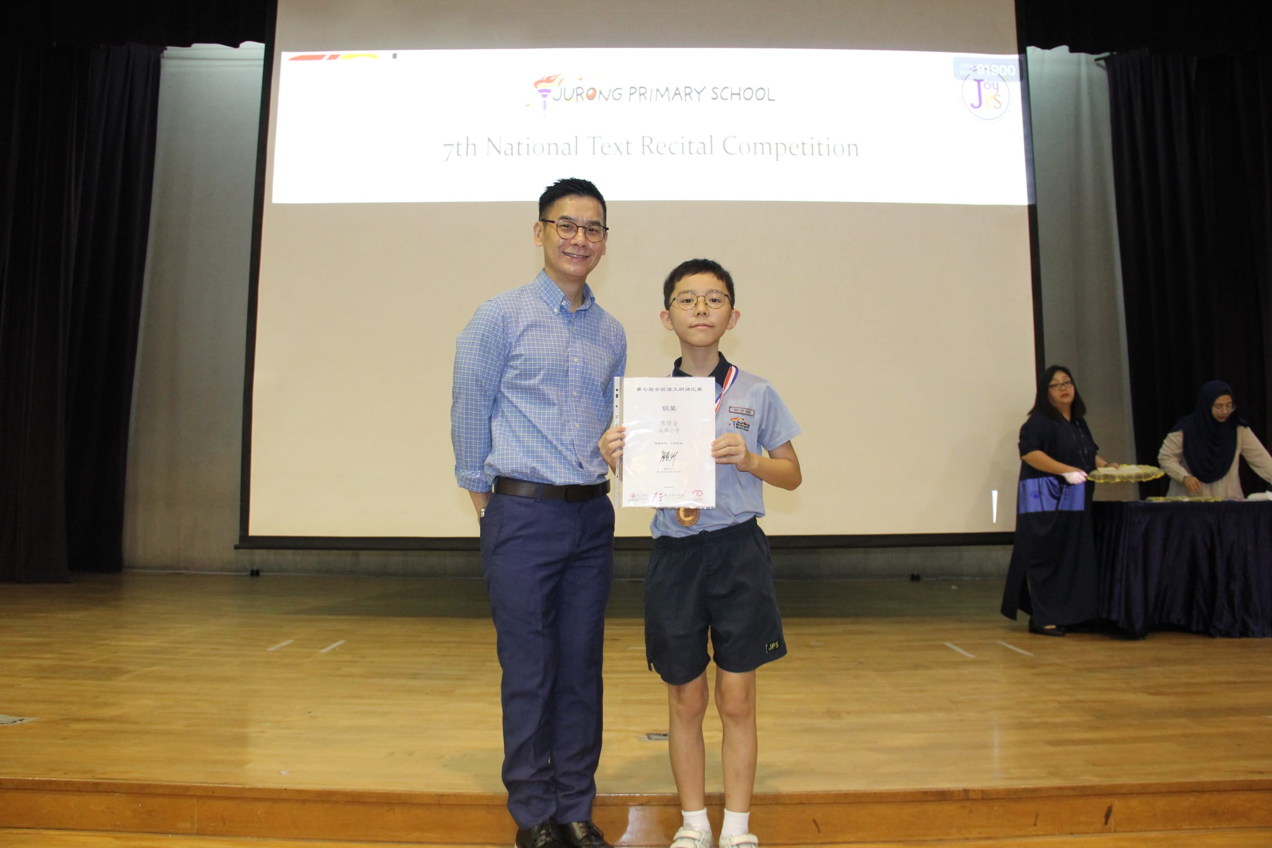 A colour photograph of a male teacher and a young student in a light blue school uniform posing together on a school stage. The student holds a certificate and wears a medal around his neck. A projection screen behind them reads "Jurong Primary School — 7th National Text Recital Competition." The student is celebrating his achievement in the Chinese text recital competition.