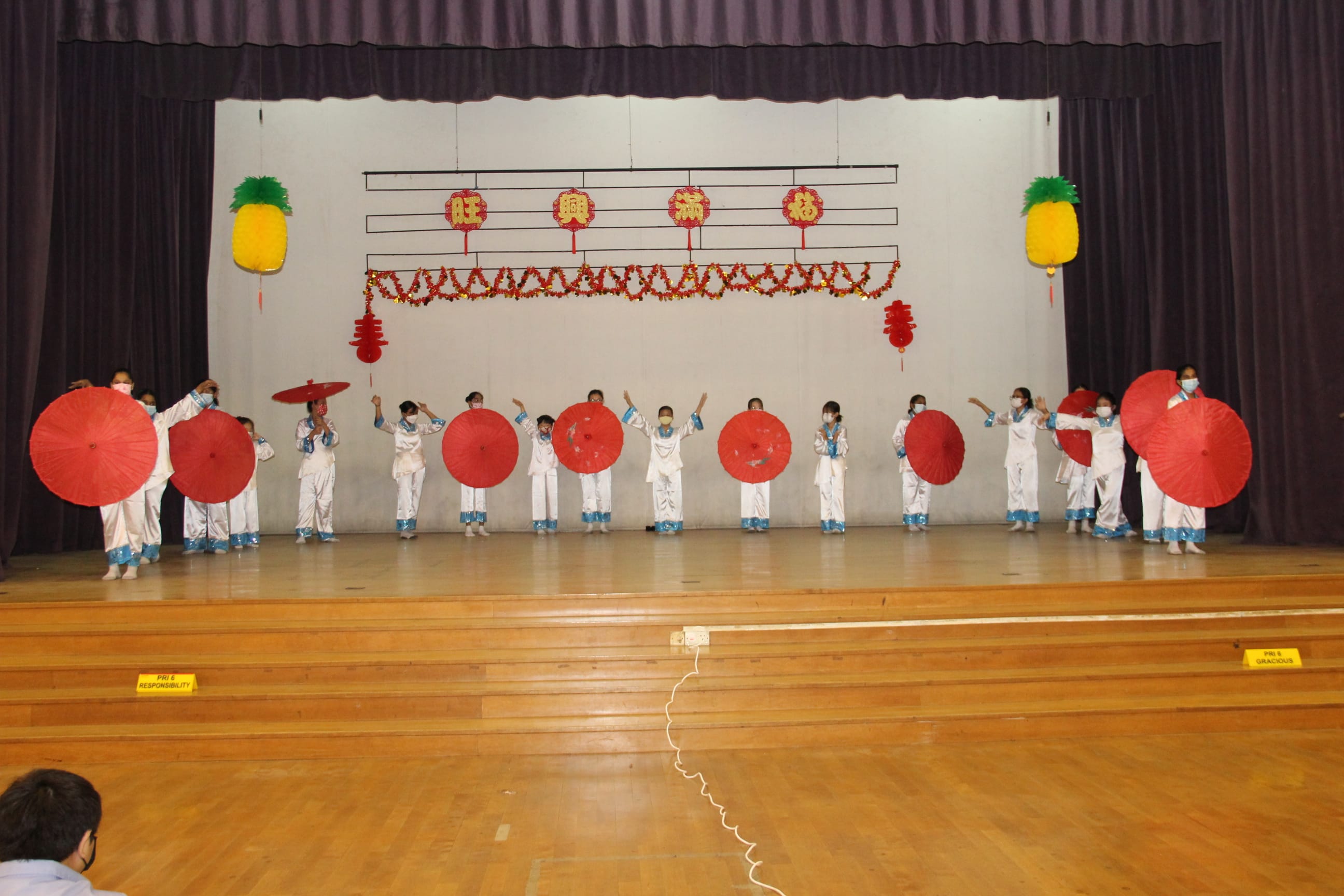 Children in white costumes hold red parasols on a stage decorated with Chinese New Year ornaments and characters.