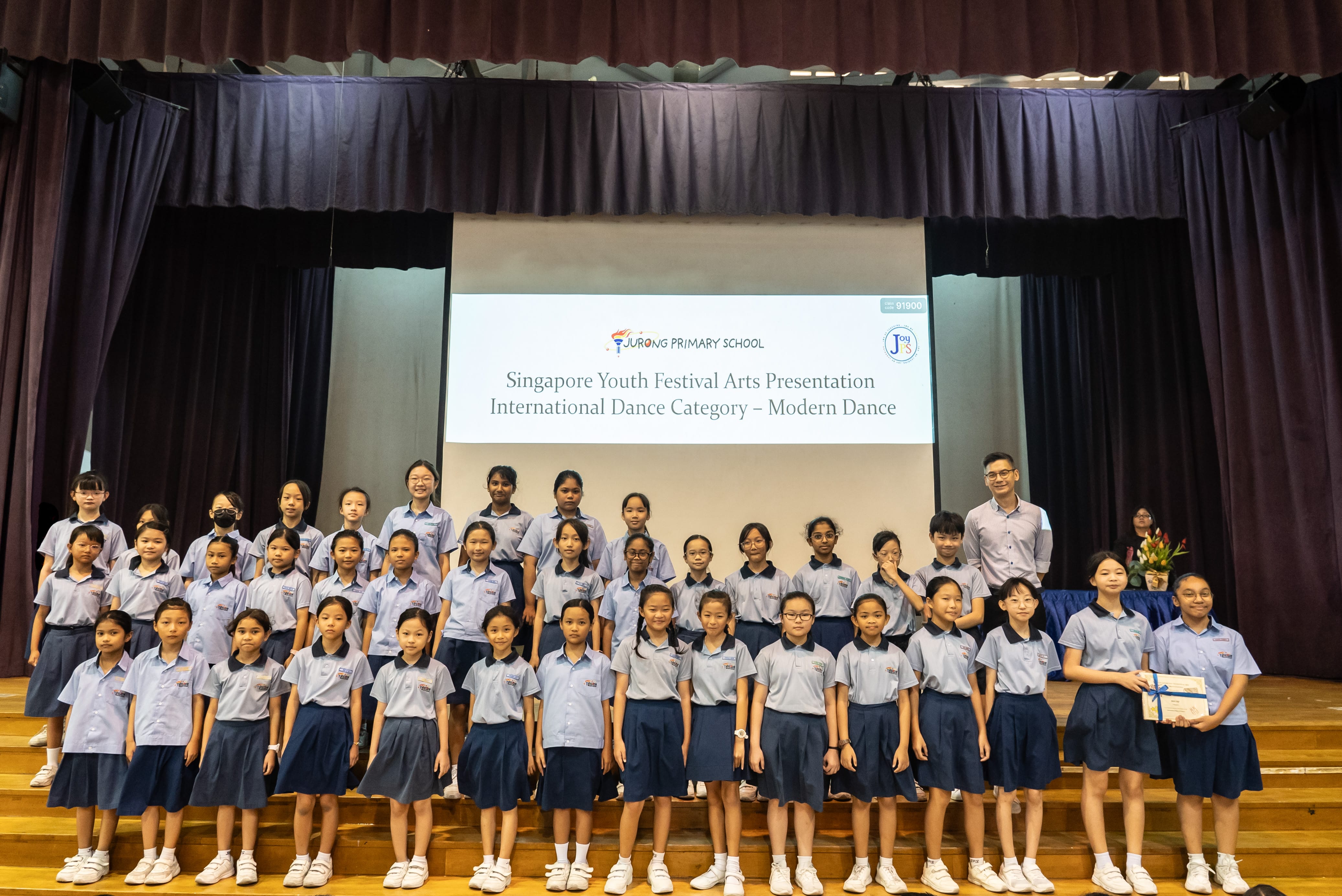 A colour photograph of a large group of approximately thirty female students in light blue school uniforms posing on a school stage with a male teacher. A projection screen behind them reads "Jurong Primary School — Singapore Youth Festival Arts Presentation, International Dance Category – Modern Dance." One student on the right holds a certificate tied with a blue ribbon. The group is celebrating their participation or achievement in the dance event.