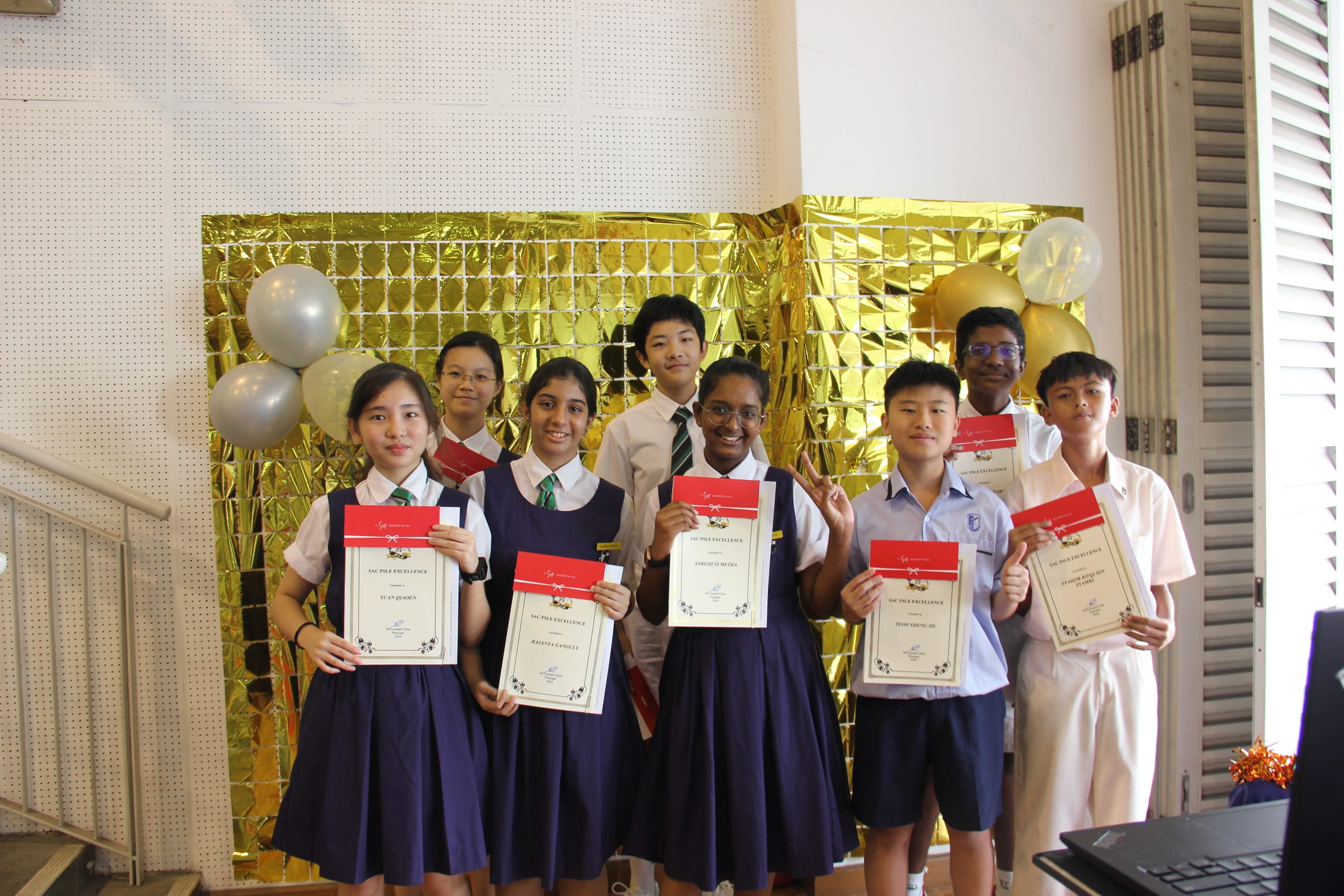 A colour photograph of eight students in school uniforms posing together in front of a shimmering gold backdrop decorated with silver and gold balloons. Each student proudly holds a certificate with a red ribbon, labelled "SAC PSLE Excellence." The students are smiling and appear to be celebrating their academic achievement. The setting appears to be indoors at a school.