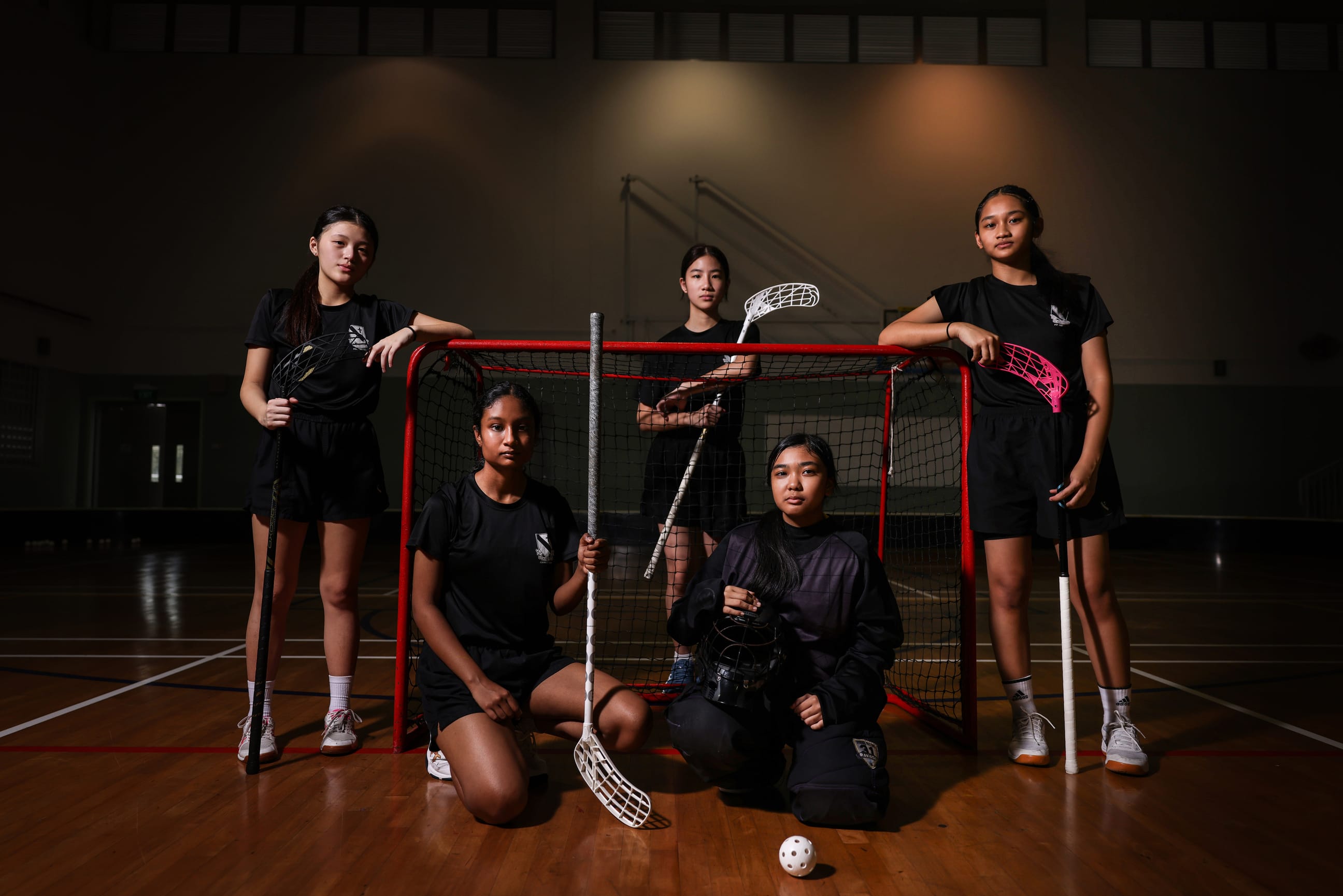 Five girls in black uniforms pose with floorball sticks around a red goal net in an indoor gymnasium.