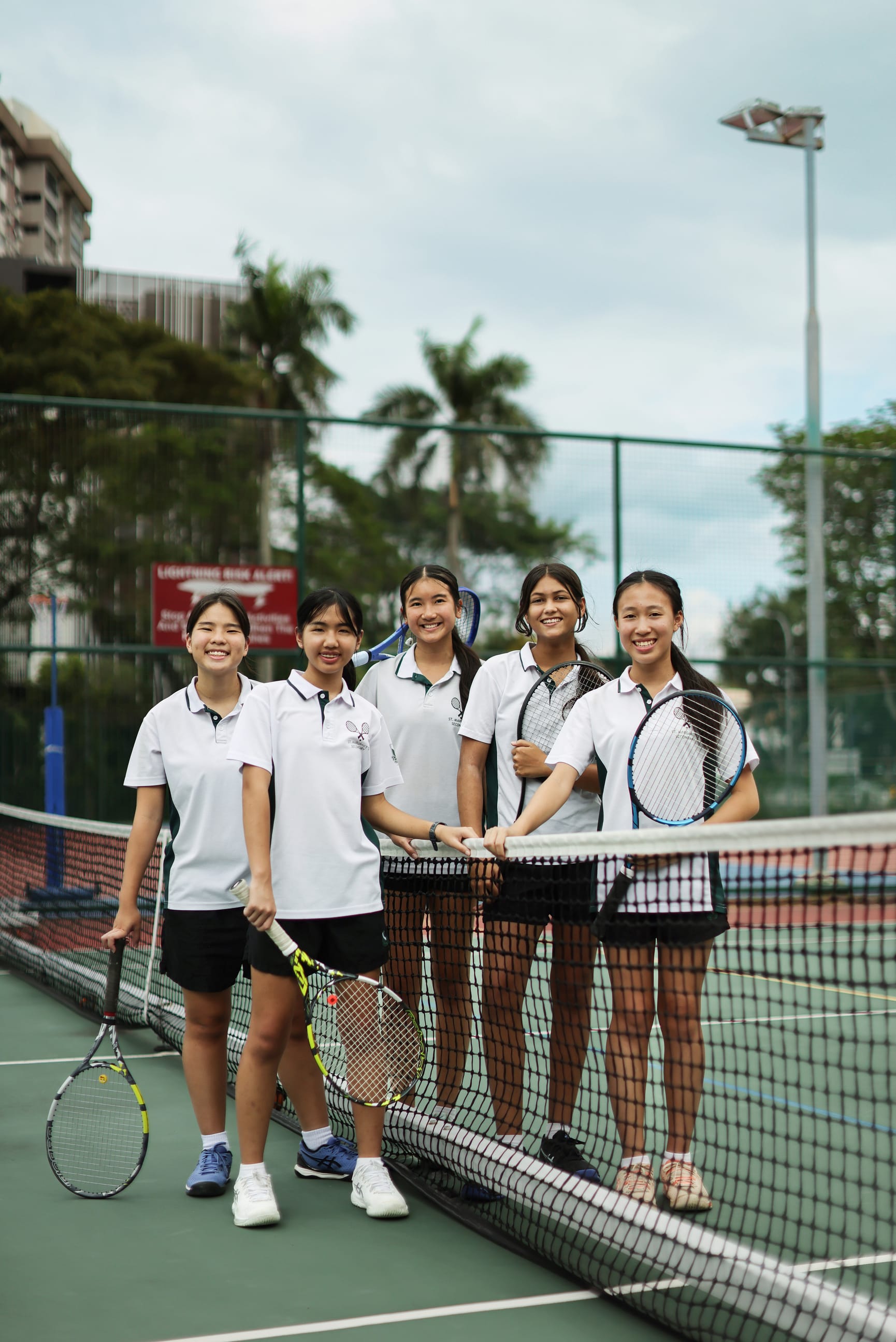 Five girls in white shirts hold tennis rackets and smile by the net on an outdoor court.