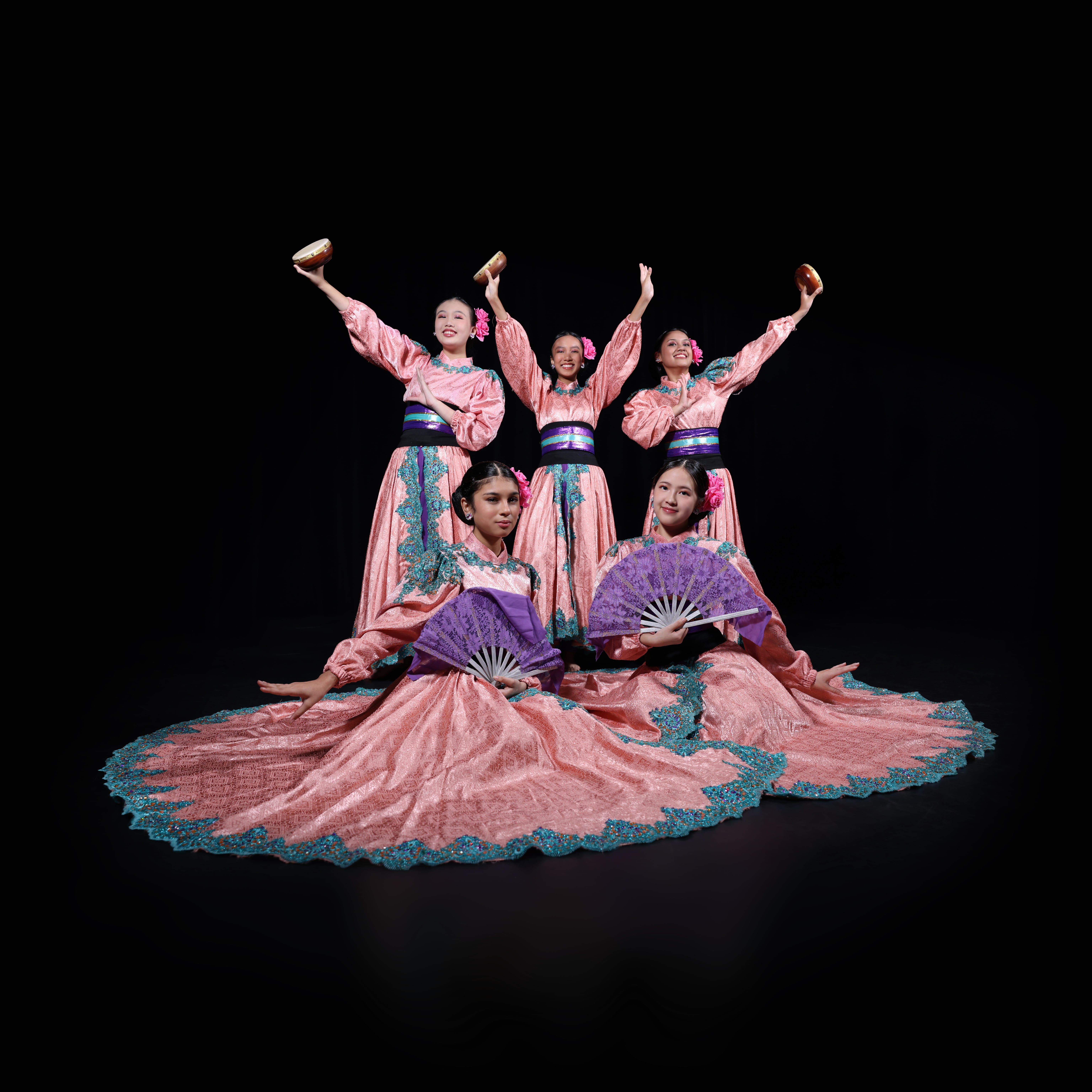 Five dancers in pink gowns; three stand with arms raised holding bowls, two sit holding purple fans.