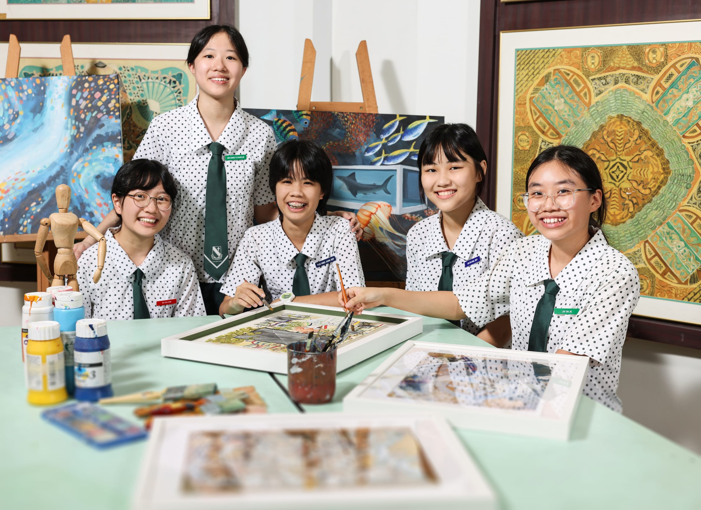 Five smiling female students in polka-dot uniforms paint at a table surrounded by colorful artwork on the walls.