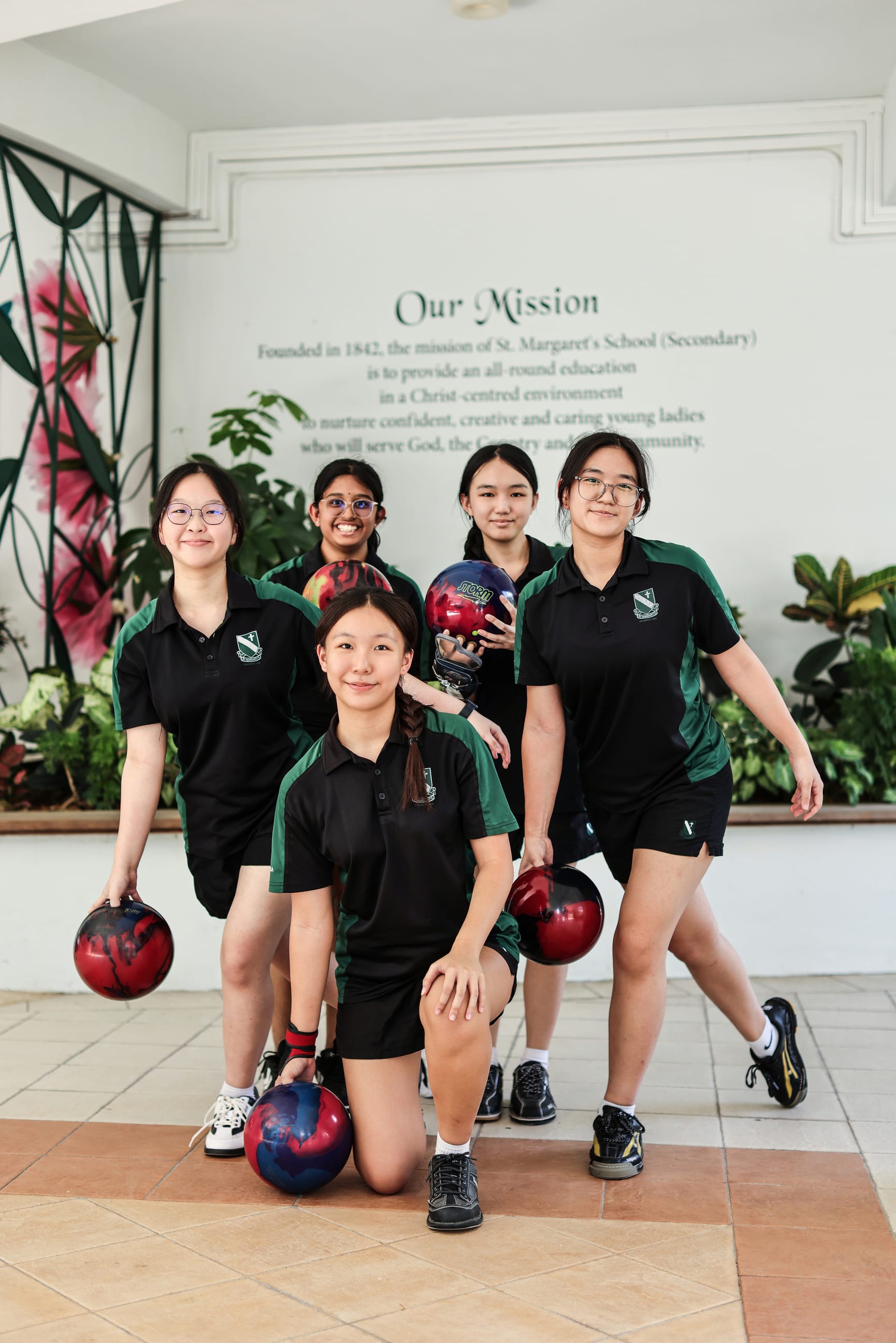Five girls in black and green school uniforms pose with bowling balls in front of a mission statement wall.