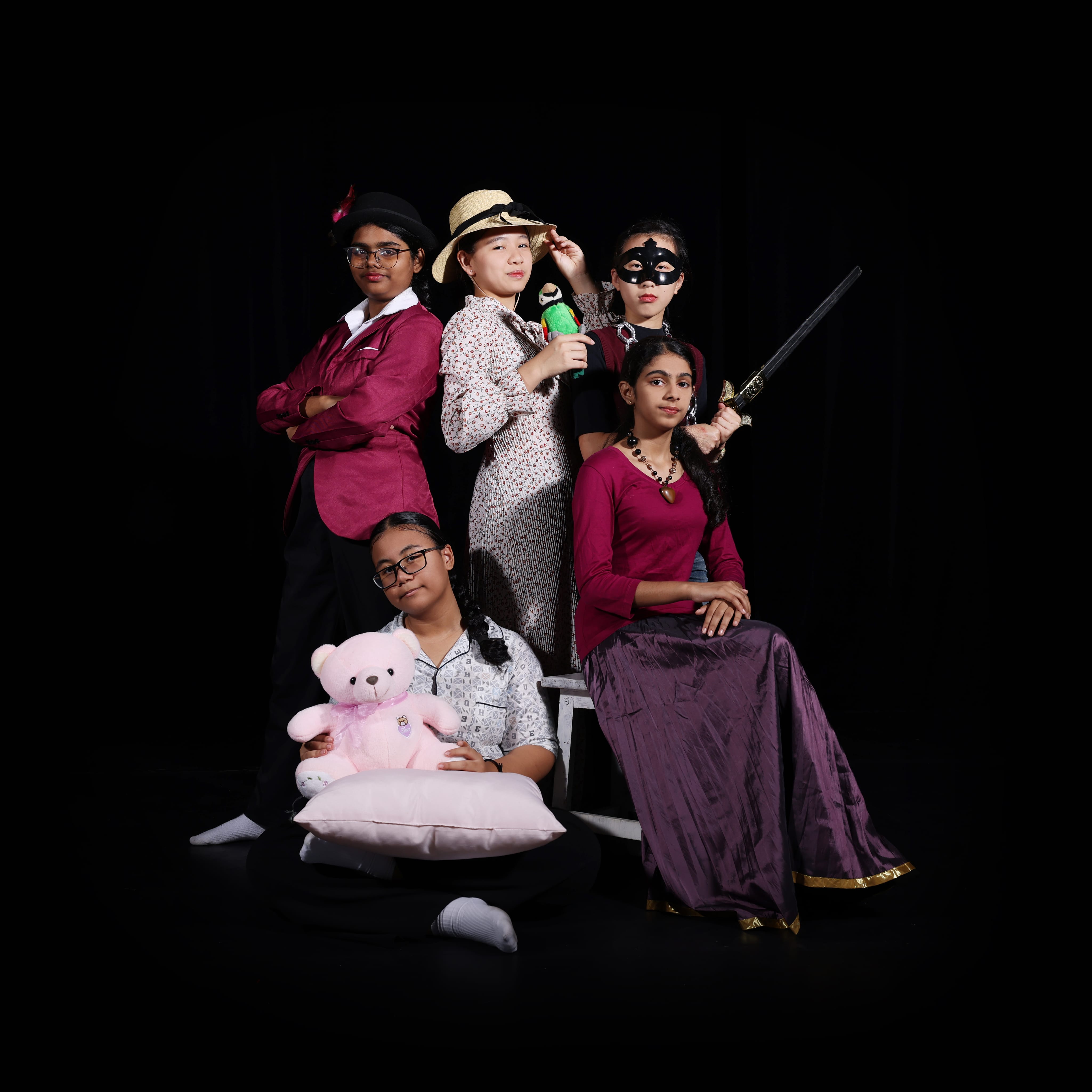 Five people pose in costumes on a black background; one holds a sword, one a toy gun, one a teddy bear.