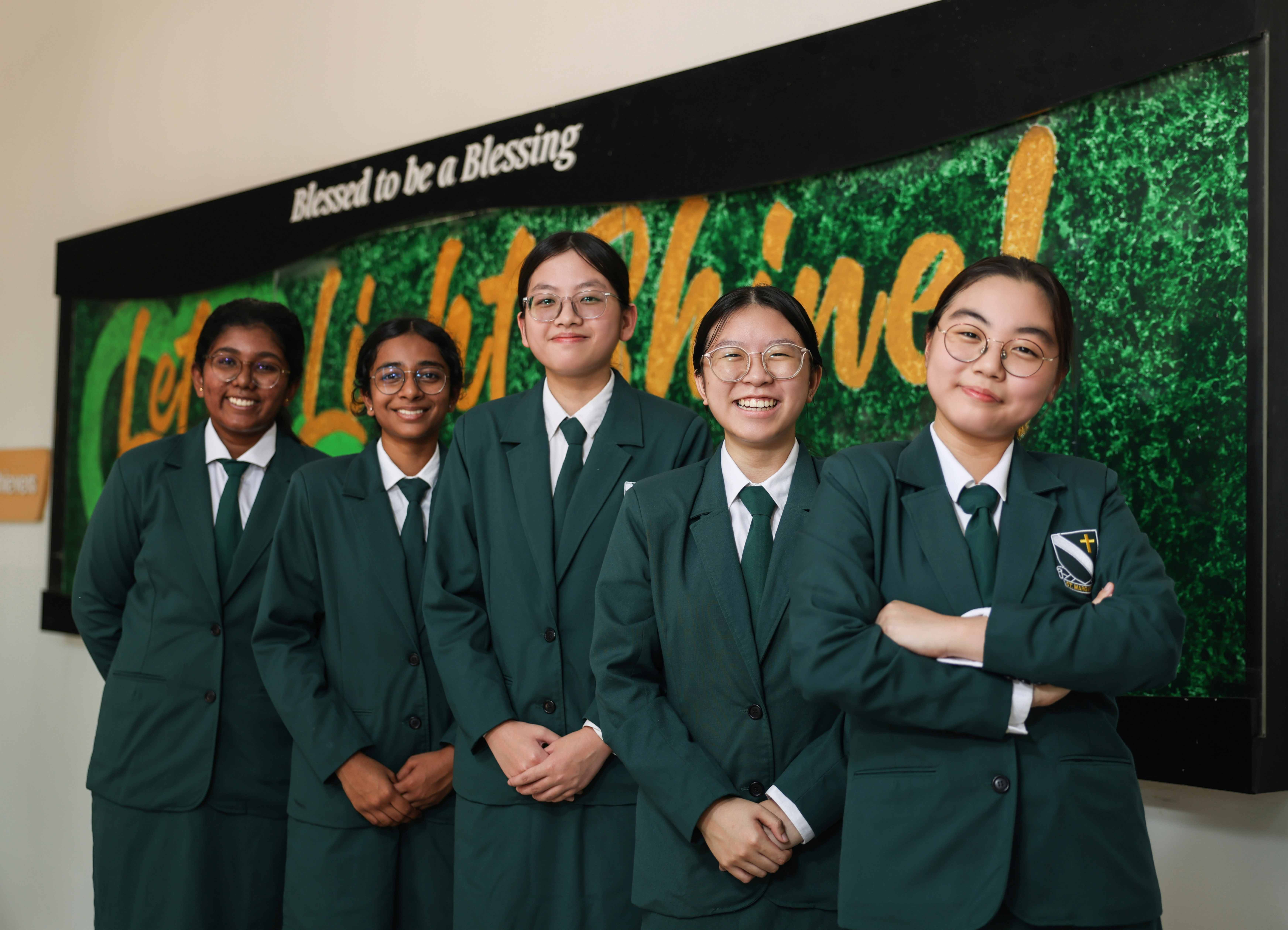 Five smiling female students in green school uniforms stand before a sign reading "Blessed to be a Blessing."